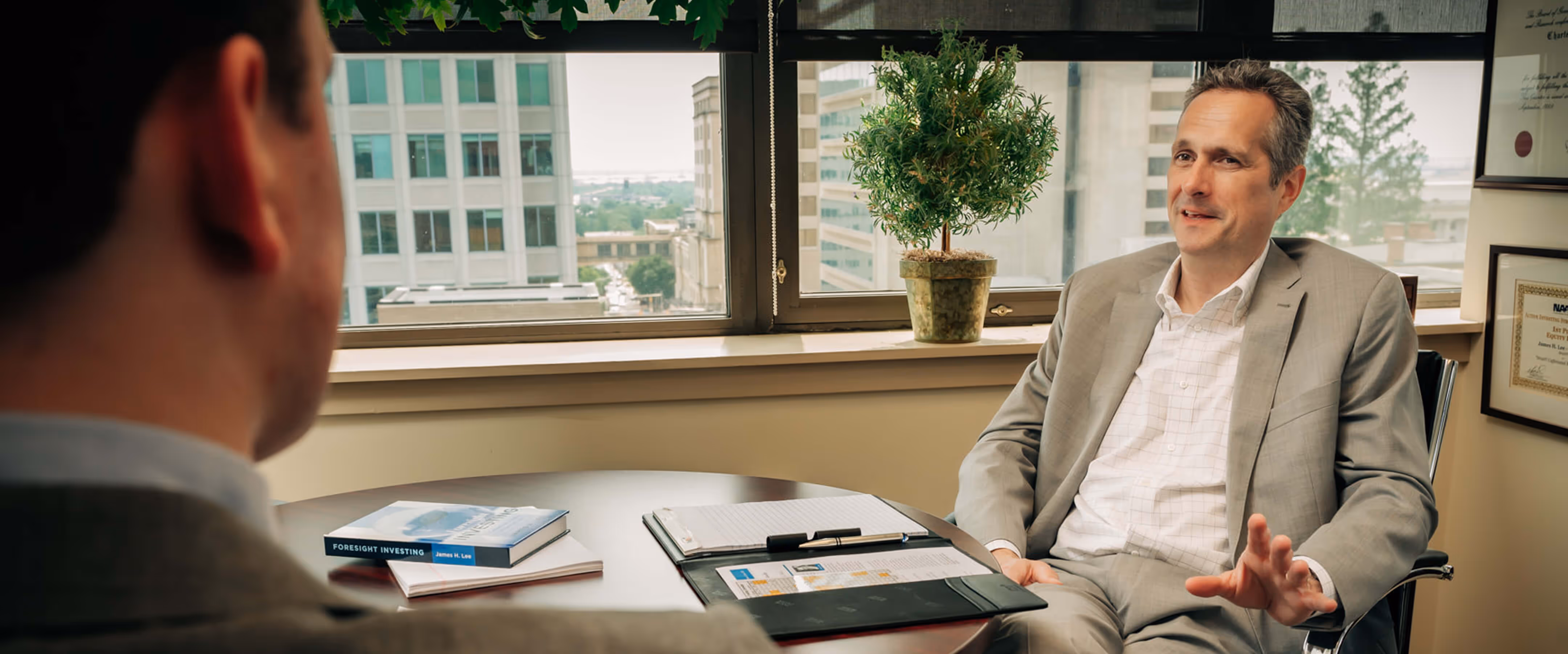 James H. Lee and a man having a conversation in an office with a round table, documents, and a plant by the window.
