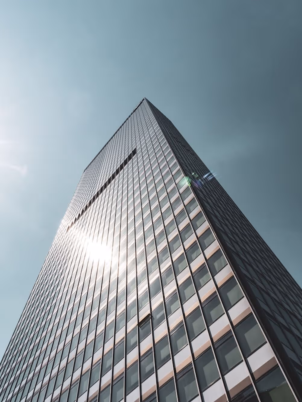 Looking up at a tall glass skyscraper reflecting sunlight against a clear blue sky.