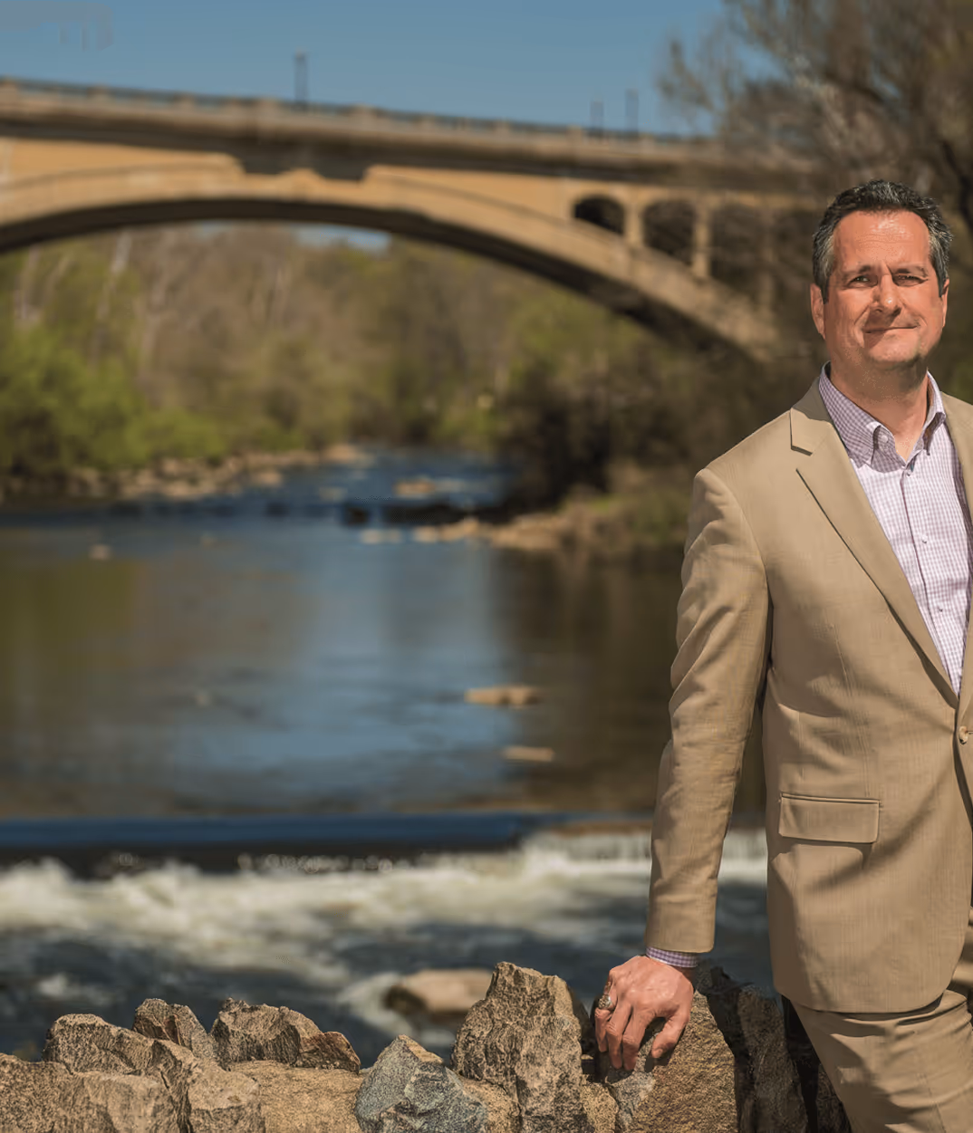 James H. Lee in tan suit standing by a stone wall with a river and arched bridge in the background.