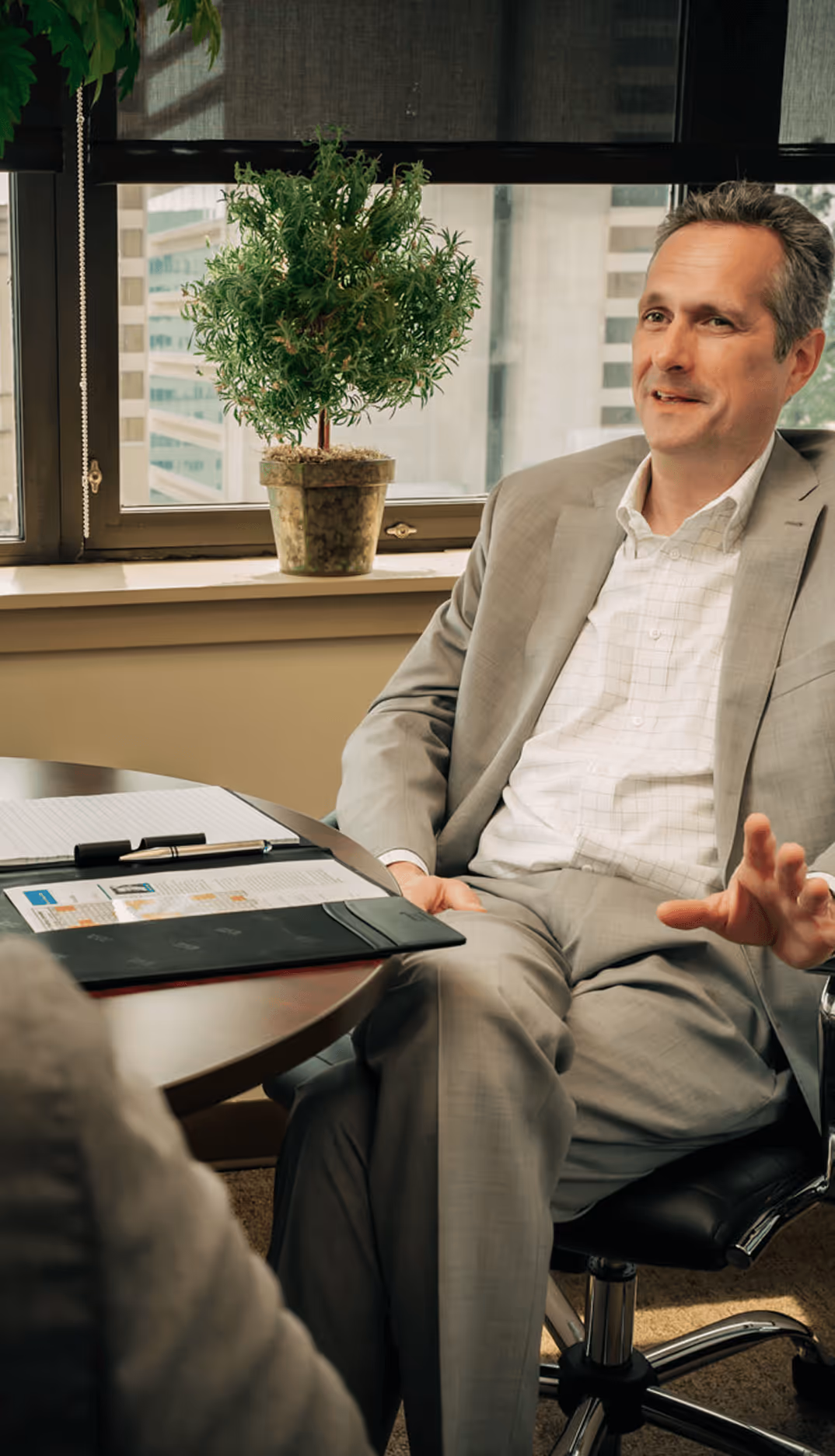 James H. Lee in a gray suit sitting on an office chair at a round table during a business meeting, with a large window and a potted green plant behind him.