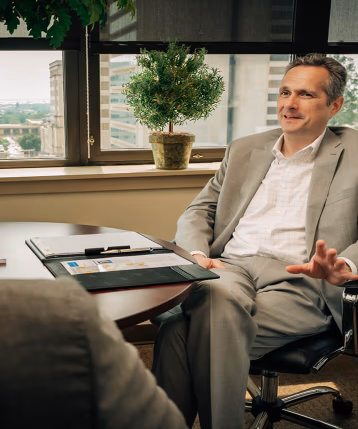 James H. Lee in a light gray suit sitting on a black office chair, talking during a meeting in an office with a round table and a potted plant on the windowsill.