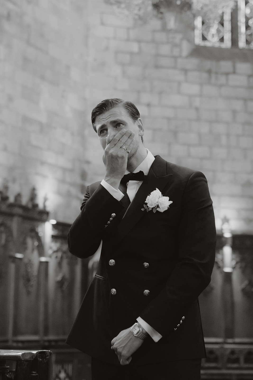 Man in formal suit with a white boutonniere covering his mouth, appearing emotional inside a stone-walled venue.