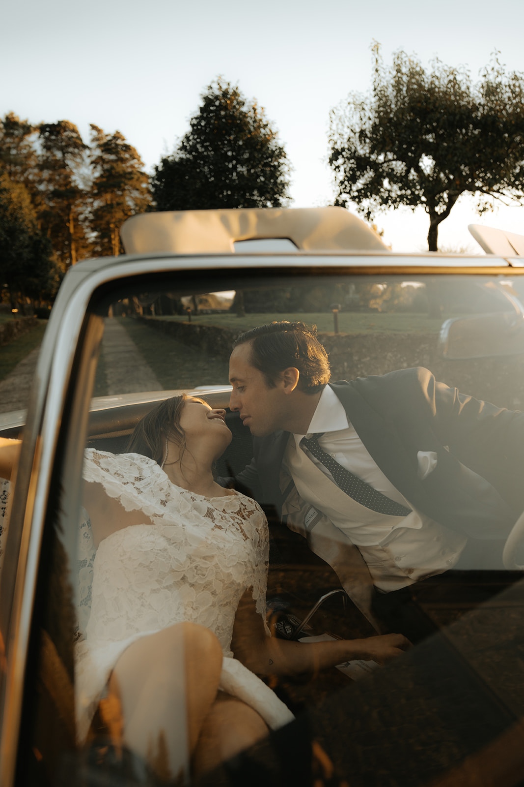 Bride in lace dress and groom in suit leaning towards each other inside a vintage convertible car at sunset.