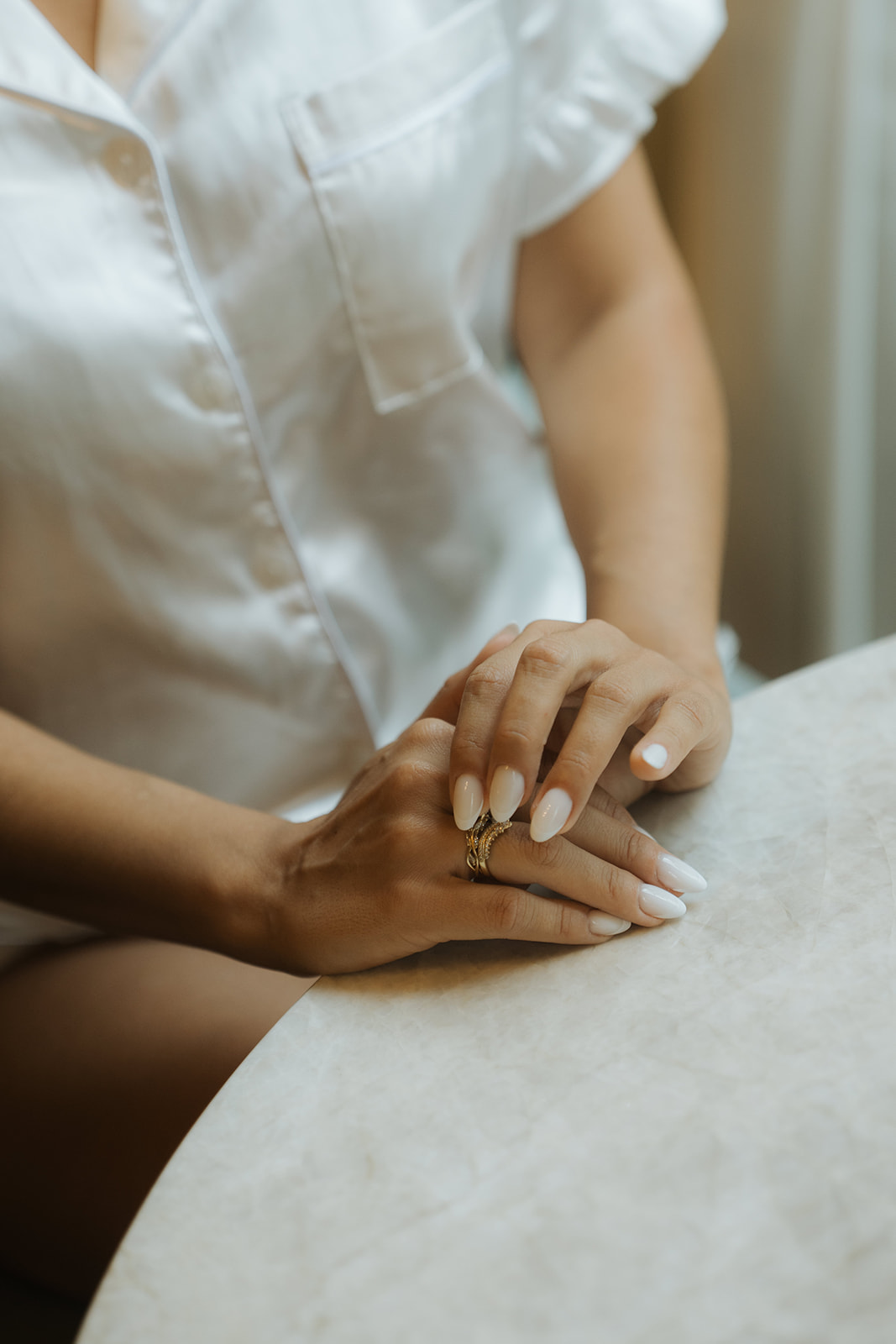 Person with manicured nails wearing gold rings resting hands on a white marble table, dressed in a white buttoned shirt.