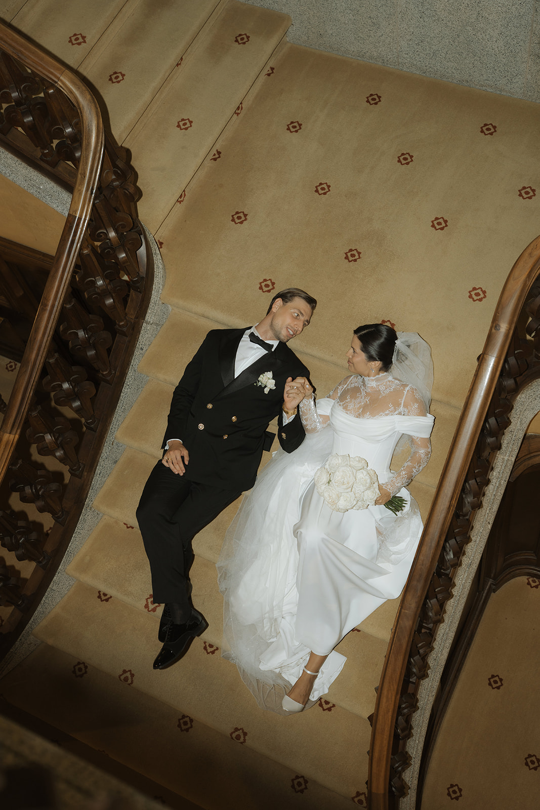 Bride in white wedding dress holding bouquet and groom in black tuxedo lying on a patterned staircase, holding hands and looking at each other.
