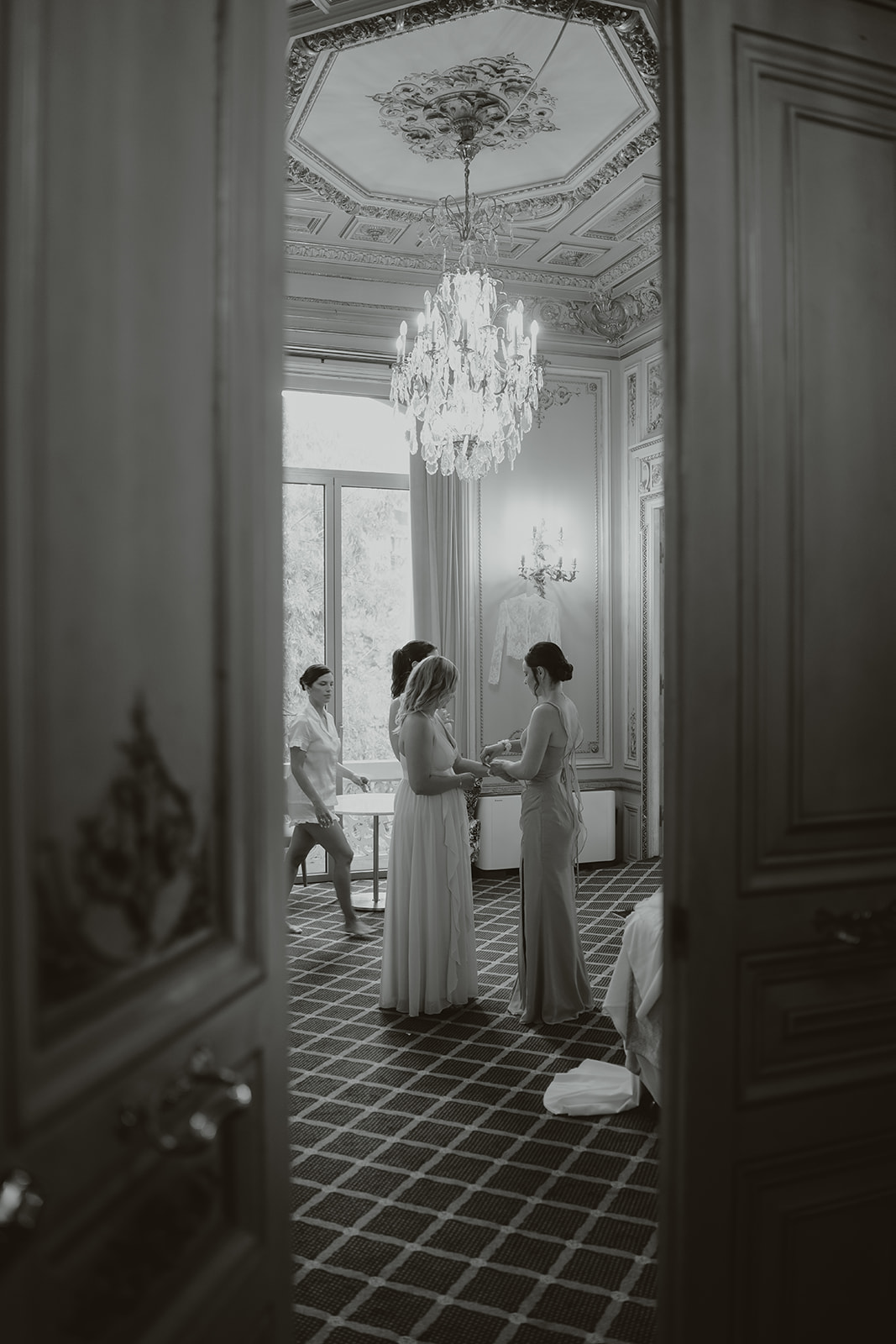 Three women in elegant dresses inside a decorated room with a chandelier, viewed through partially open doors.