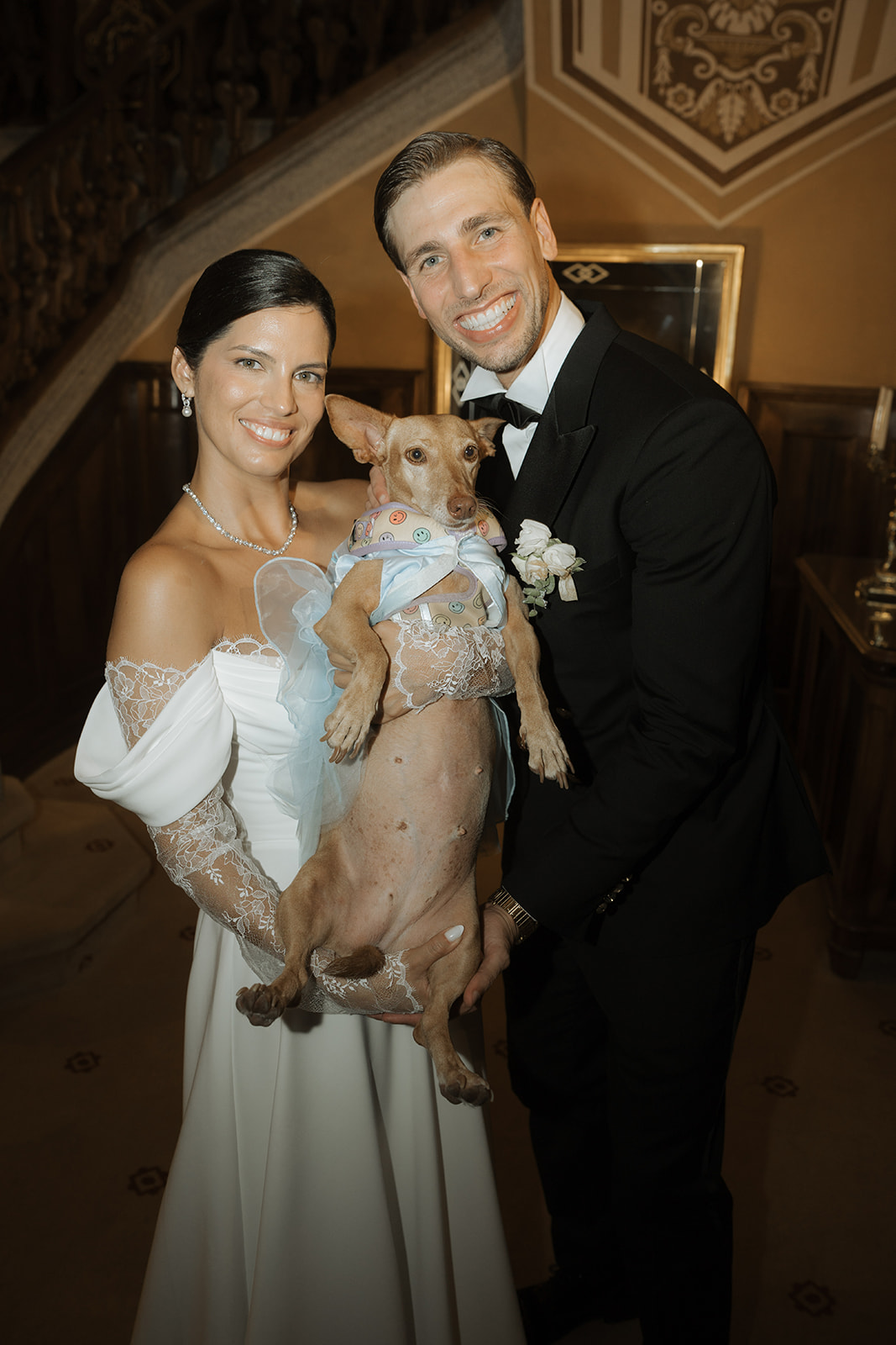 Bride and groom smiling while holding a dog dressed in a blue outfit with a smiley face collar at an indoor wedding venue.