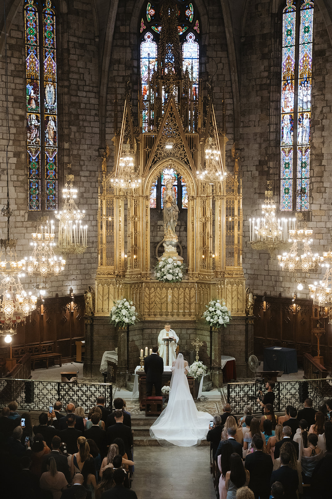 Bride and groom standing before a priest in a grand church with ornate golden altar and stained glass windows during a wedding ceremony.