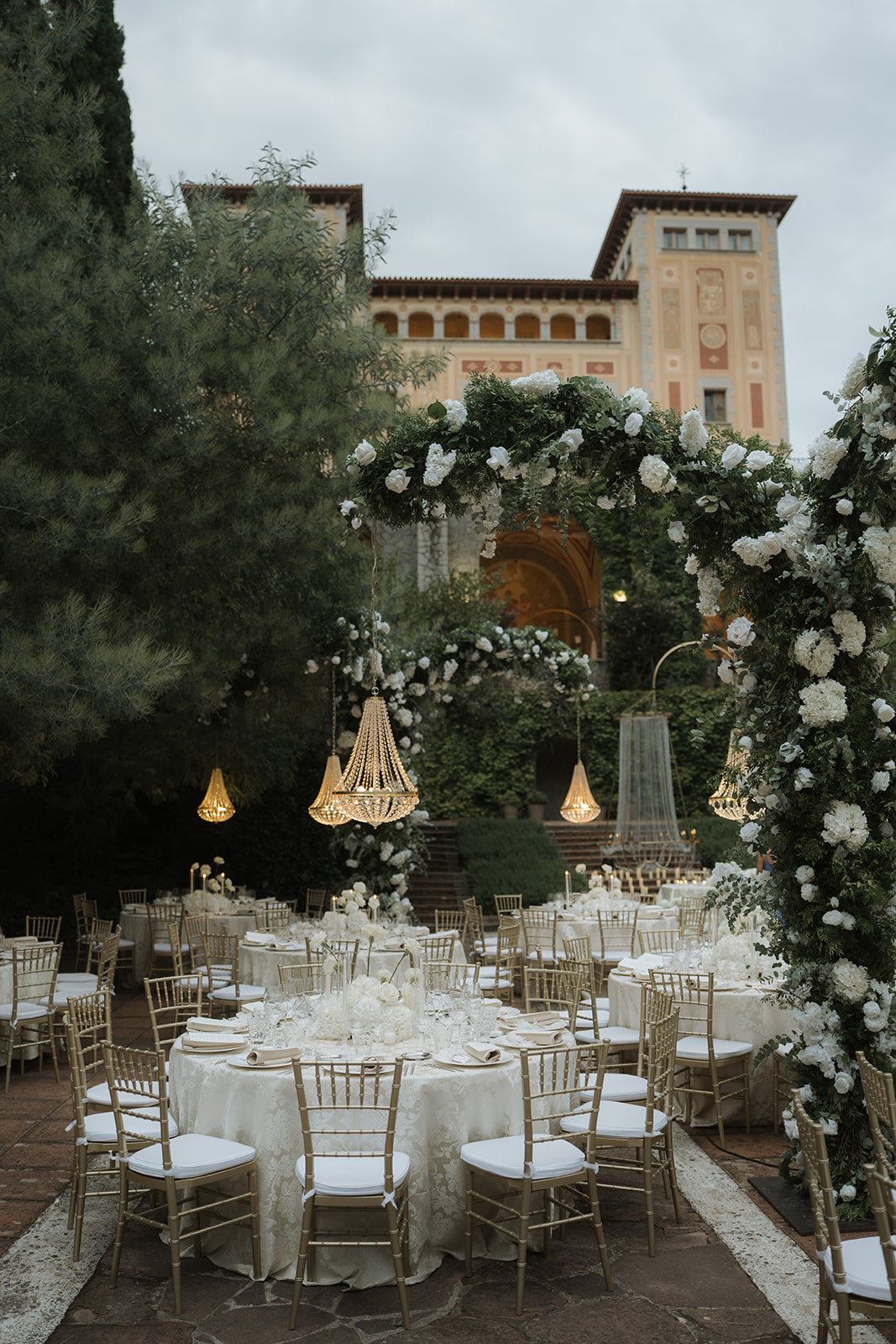 Elegant outdoor wedding setup with round tables, gold chairs, white floral arrangements, and hanging crystal chandeliers.