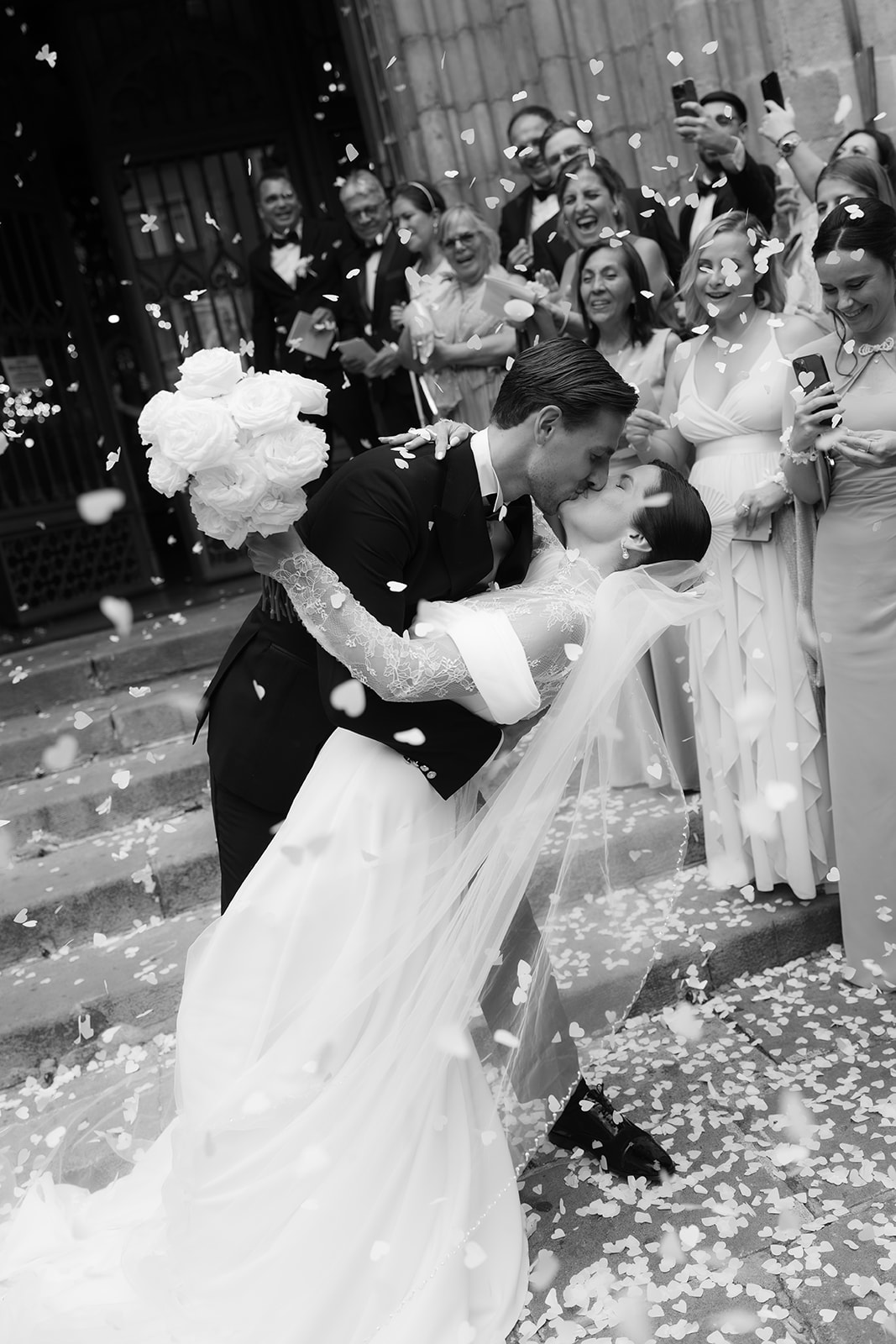 Bride and groom kissing on steps with bride holding bouquet as guests throw confetti and take photos.