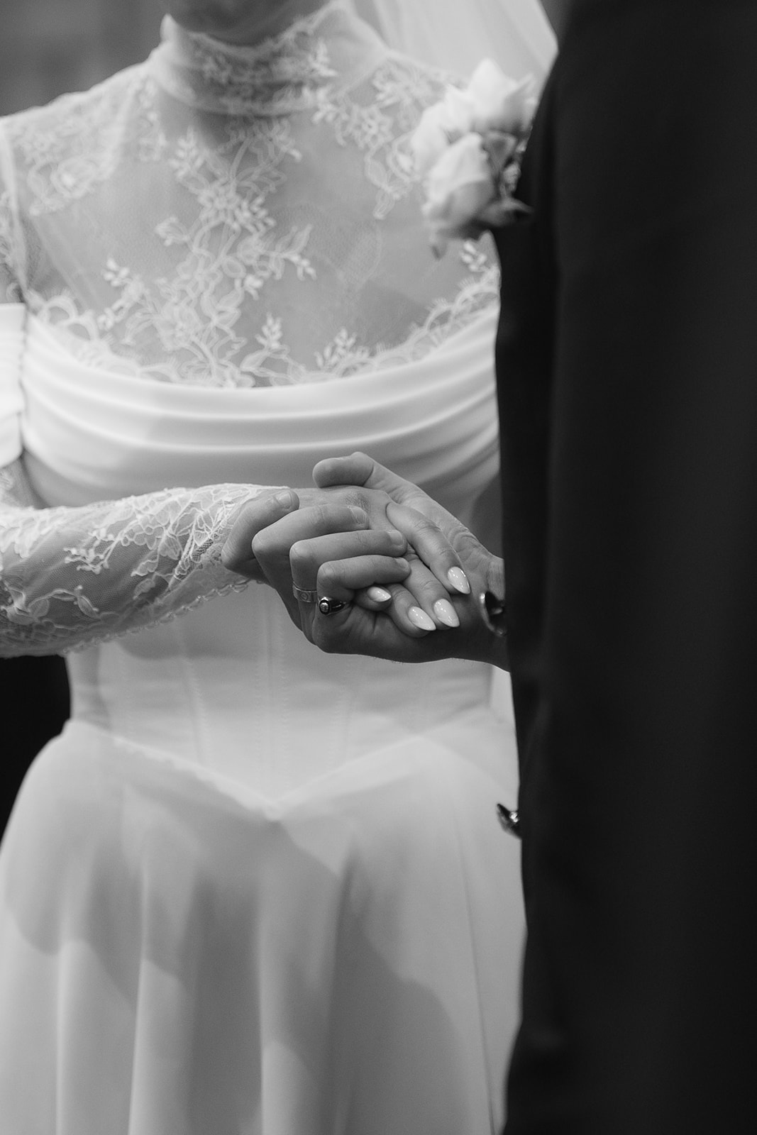 Bride and groom holding hands during wedding ceremony, bride wearing lace dress and groom in dark suit.