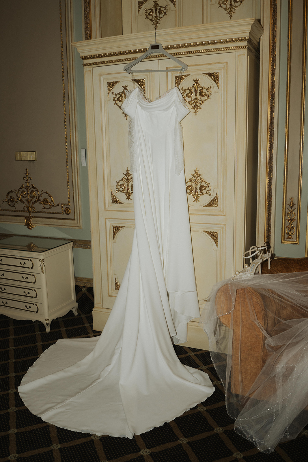 Elegant white off-shoulder wedding dress hanging on ornate cream and gold wardrobe with matching heels and veil on nearby chair.