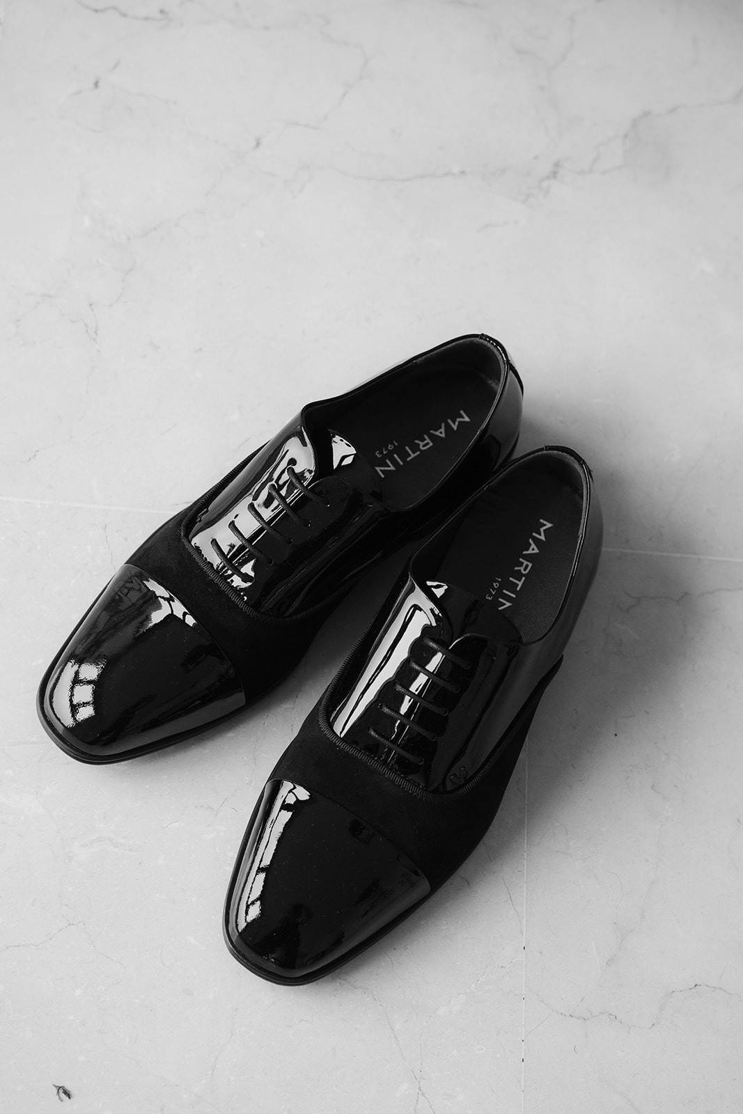 Pair of black patent leather and suede oxford dress shoes on a light marble floor.
