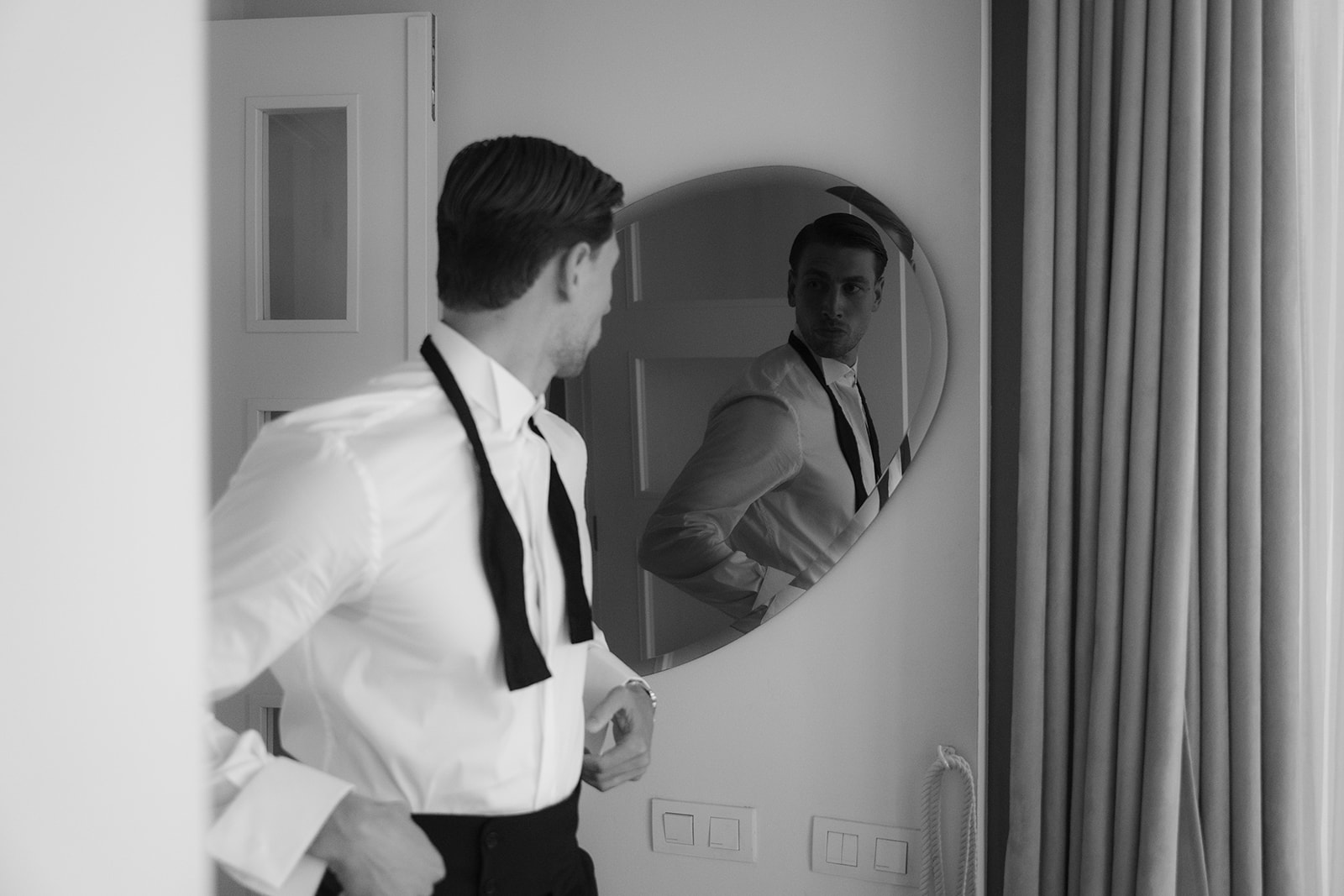 Black and white photo of a man adjusting his formal shirt and bow tie while looking at his reflection in an oval mirror.