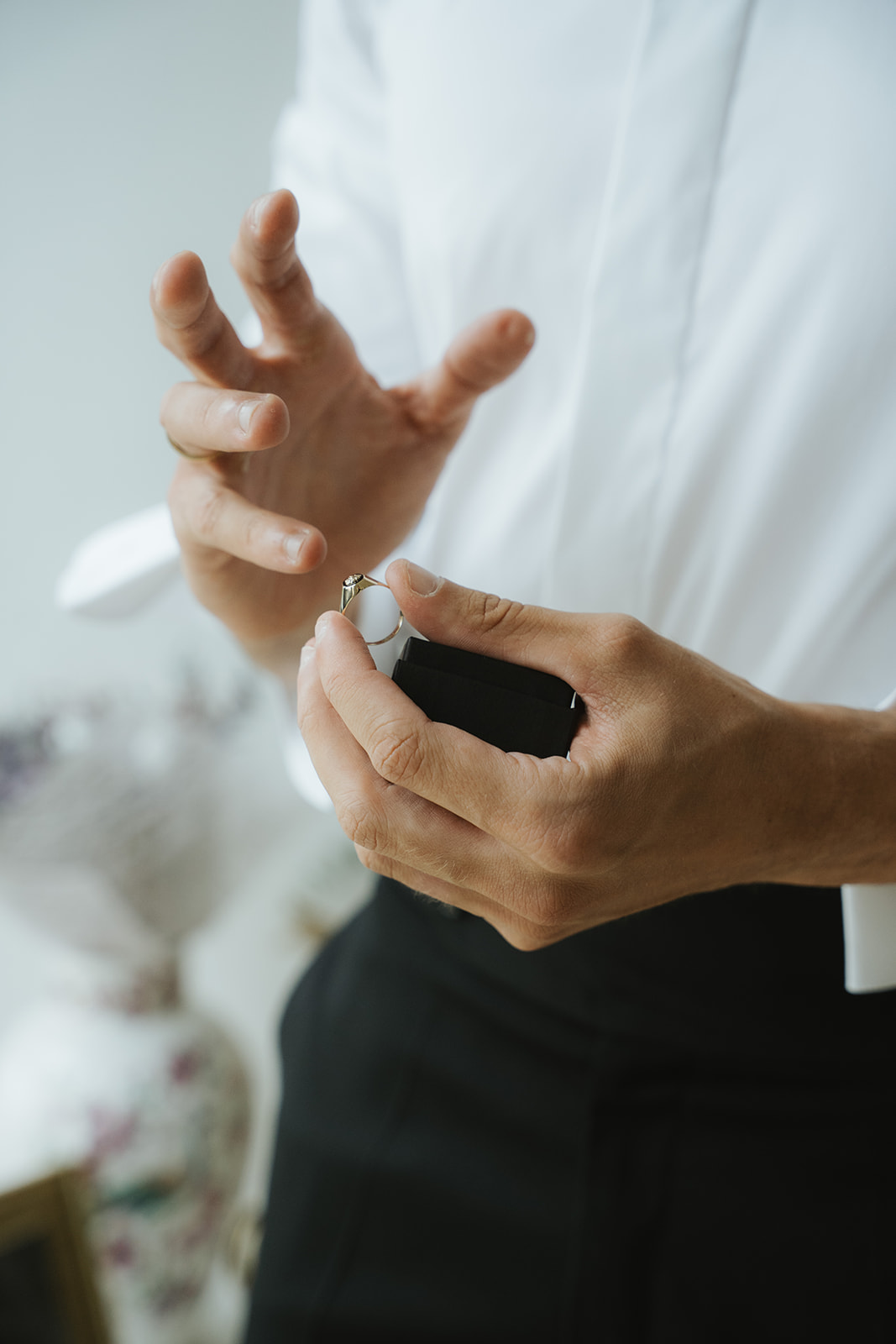 Person holding a gold engagement ring above a black ring box, wearing a white shirt and black pants.