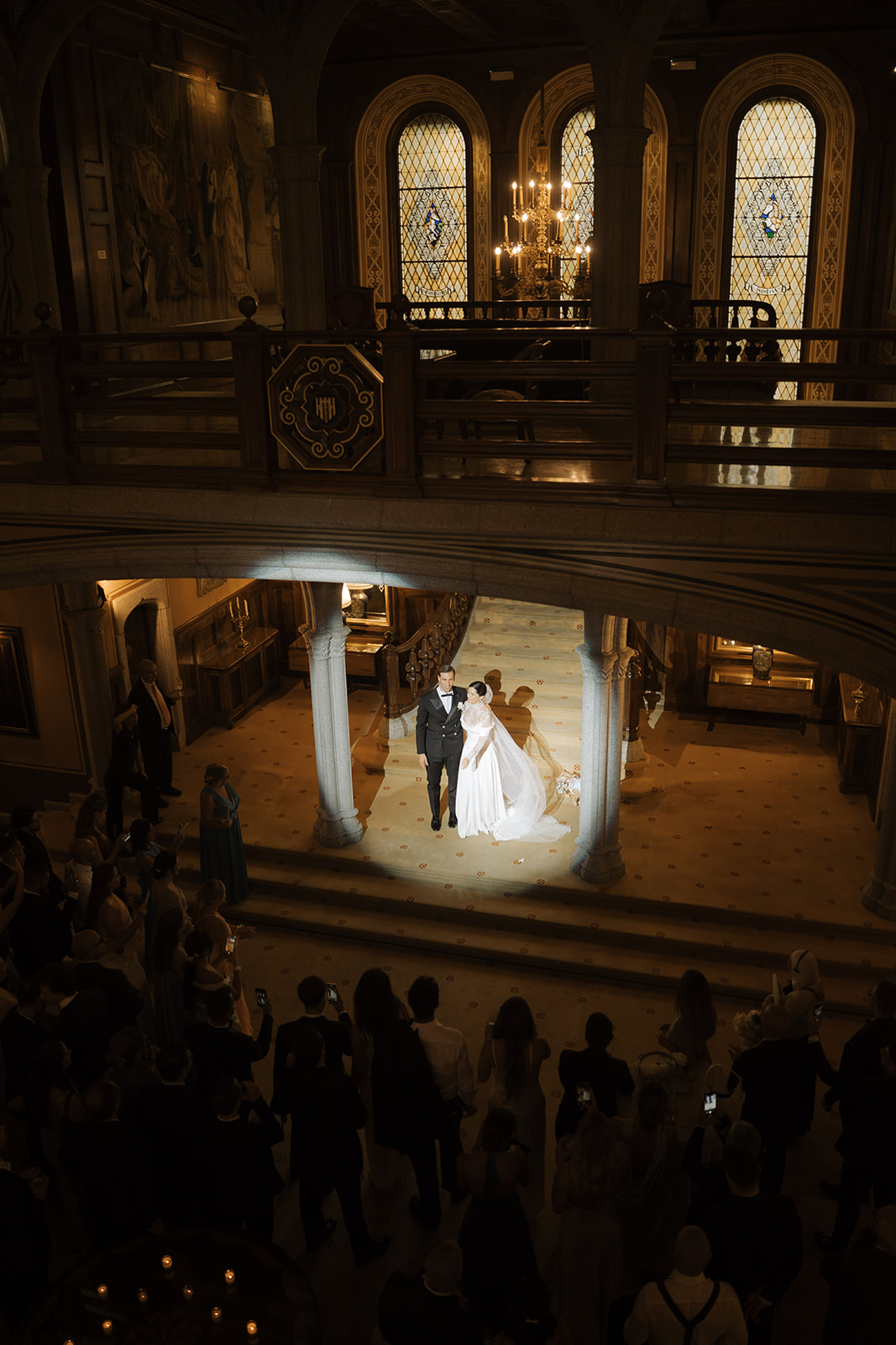 Bride and groom standing under spotlight in a grand hall surrounded by guests taking photos.