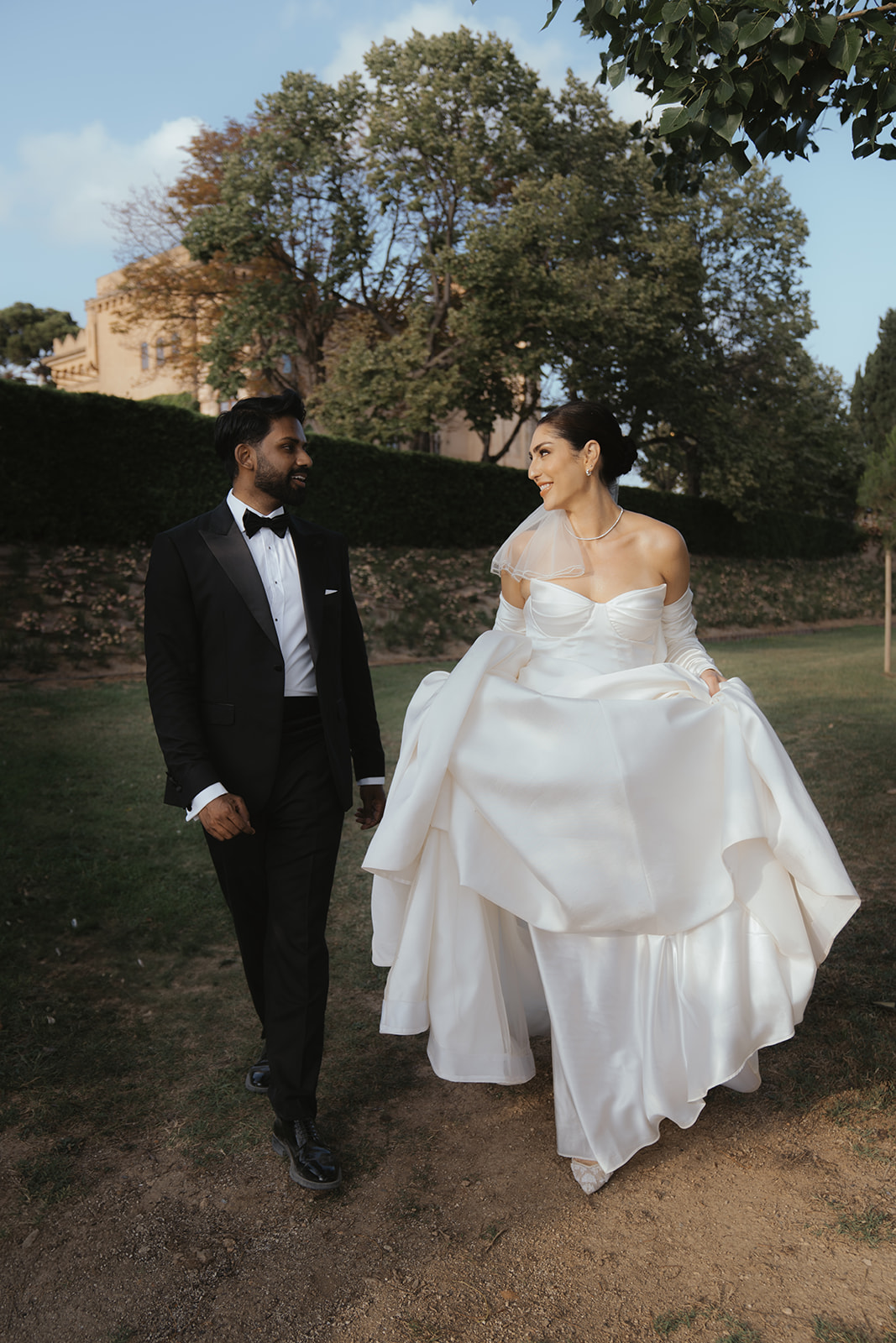 Bride in white wedding gown and groom in black tuxedo walking outdoors on a grassy path, smiling at each other.