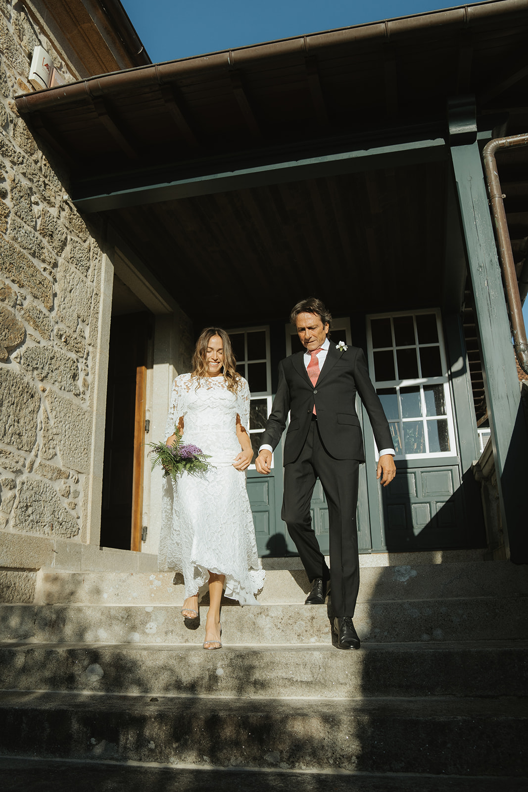 Bride and groom holding hands and smiling while walking down ornate staircase with stained glass windows and statue in the background.