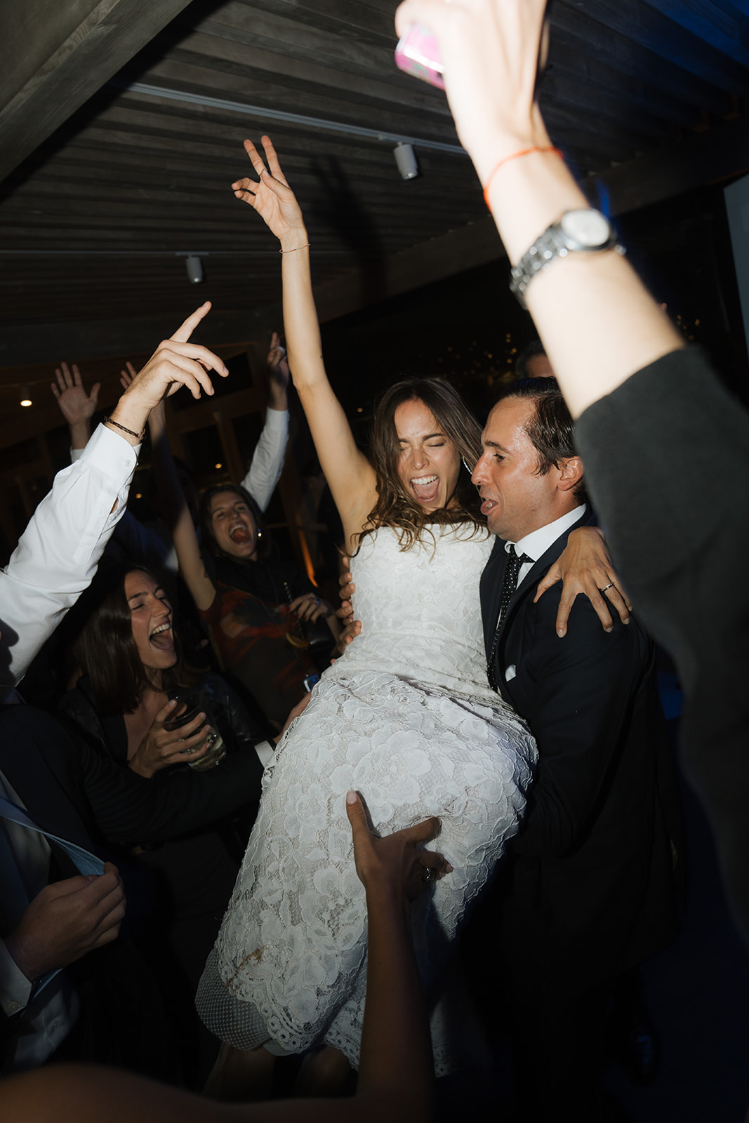 Bride and groom standing under spotlight in a grand hall surrounded by guests taking photos.