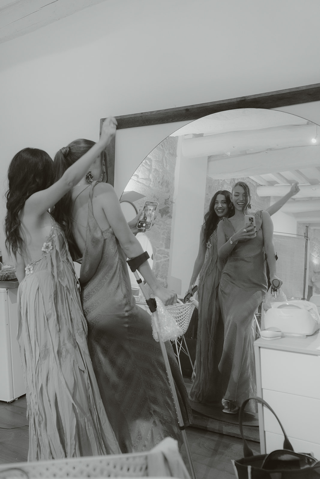 Four women in satin robes smiling and holding hands in an ornately decorated room with a wedding dress hanging in the background.