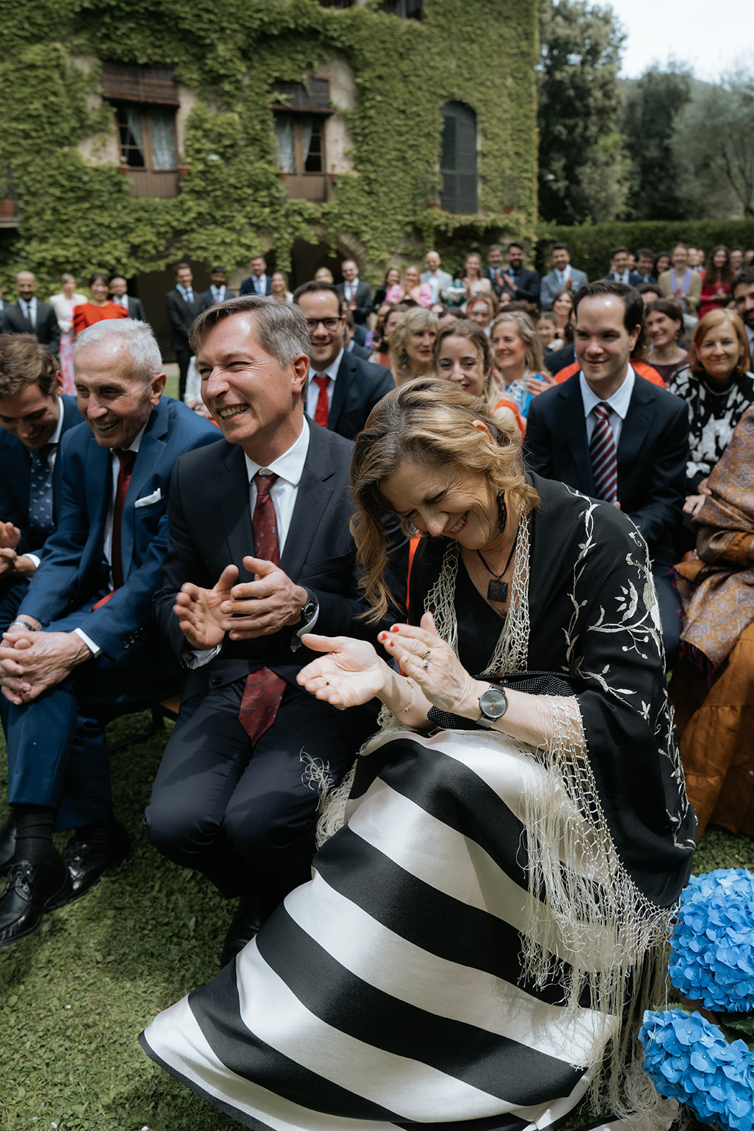 Bride and groom smiling while holding a dog dressed in a blue outfit with a smiley face collar at an indoor wedding venue.