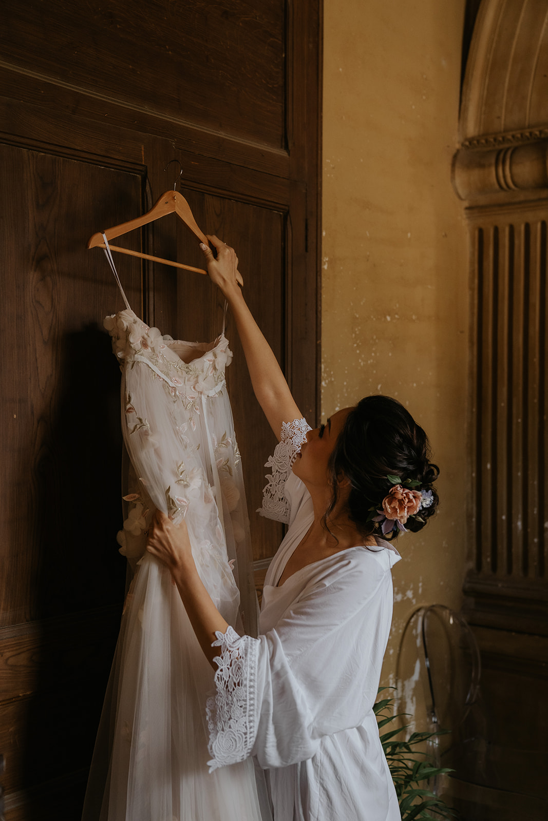 Elegant white off-shoulder wedding dress hanging on ornate cream and gold wardrobe with matching heels and veil on nearby chair.