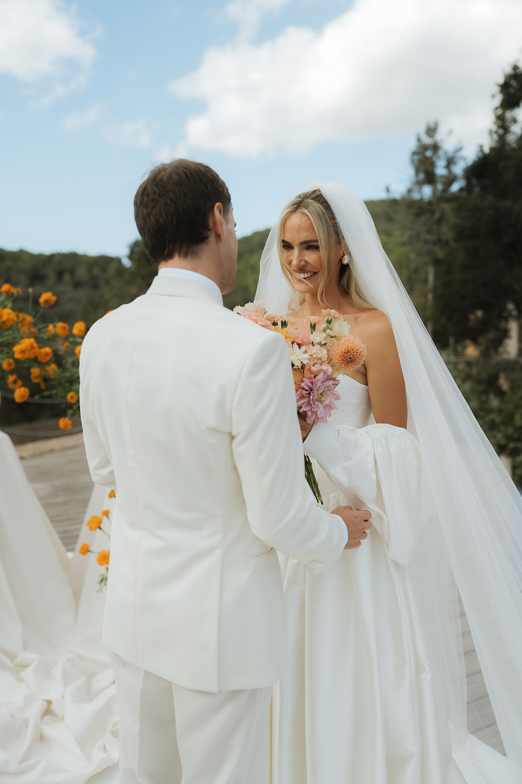 Bride in white wedding dress holding bouquet with bridesmaids in floral dresses holding her long lace veil in a vintage room.