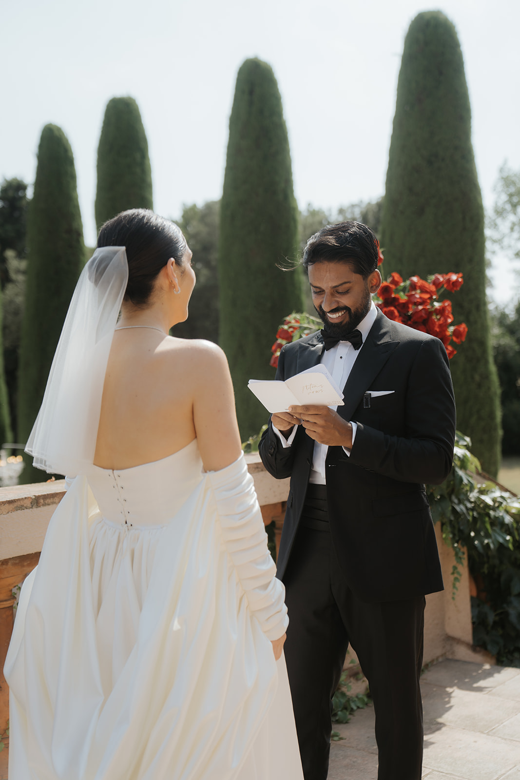 Bride and groom smiling while holding a dog dressed in a blue outfit with a smiley face collar at an indoor wedding venue.