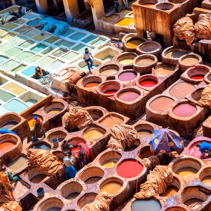 Artisanal leather tannery with workers standing and handling hides in large stone dyeing vats filled with colorful liquids.