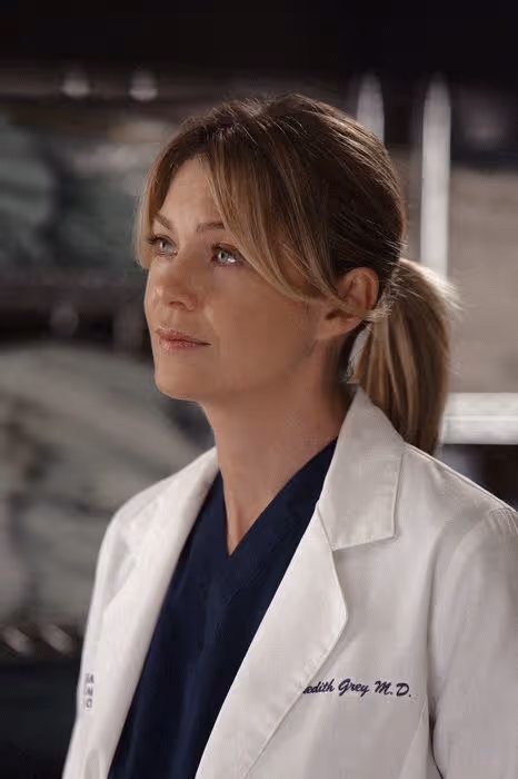 Woman with blonde hair tied back, wearing a white medical coat and navy scrubs, looking to the side.
