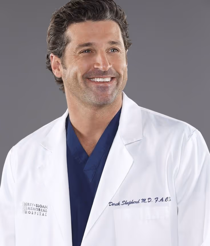 Smiling man with dark curly hair wearing a white lab coat over navy scrubs against a gray background.