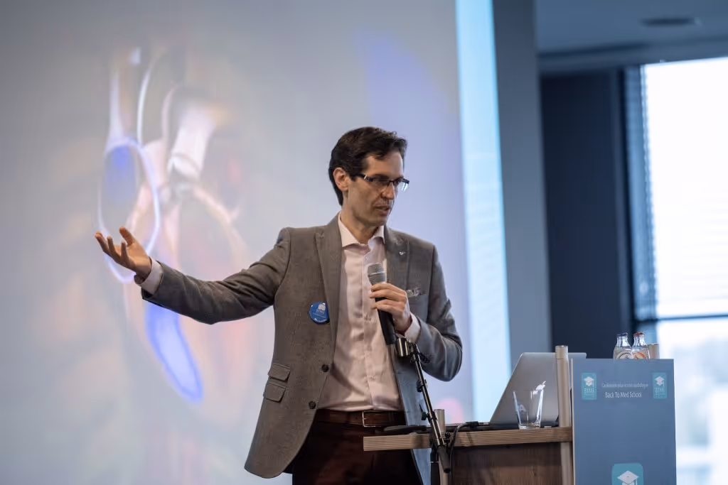 Man wearing a white lab coat and a blue checkered tie smiling with arms crossed against a plain background.