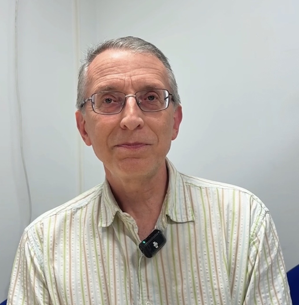 Smiling man with curly brown hair, glasses, and light beard wearing a light purple collared shirt.