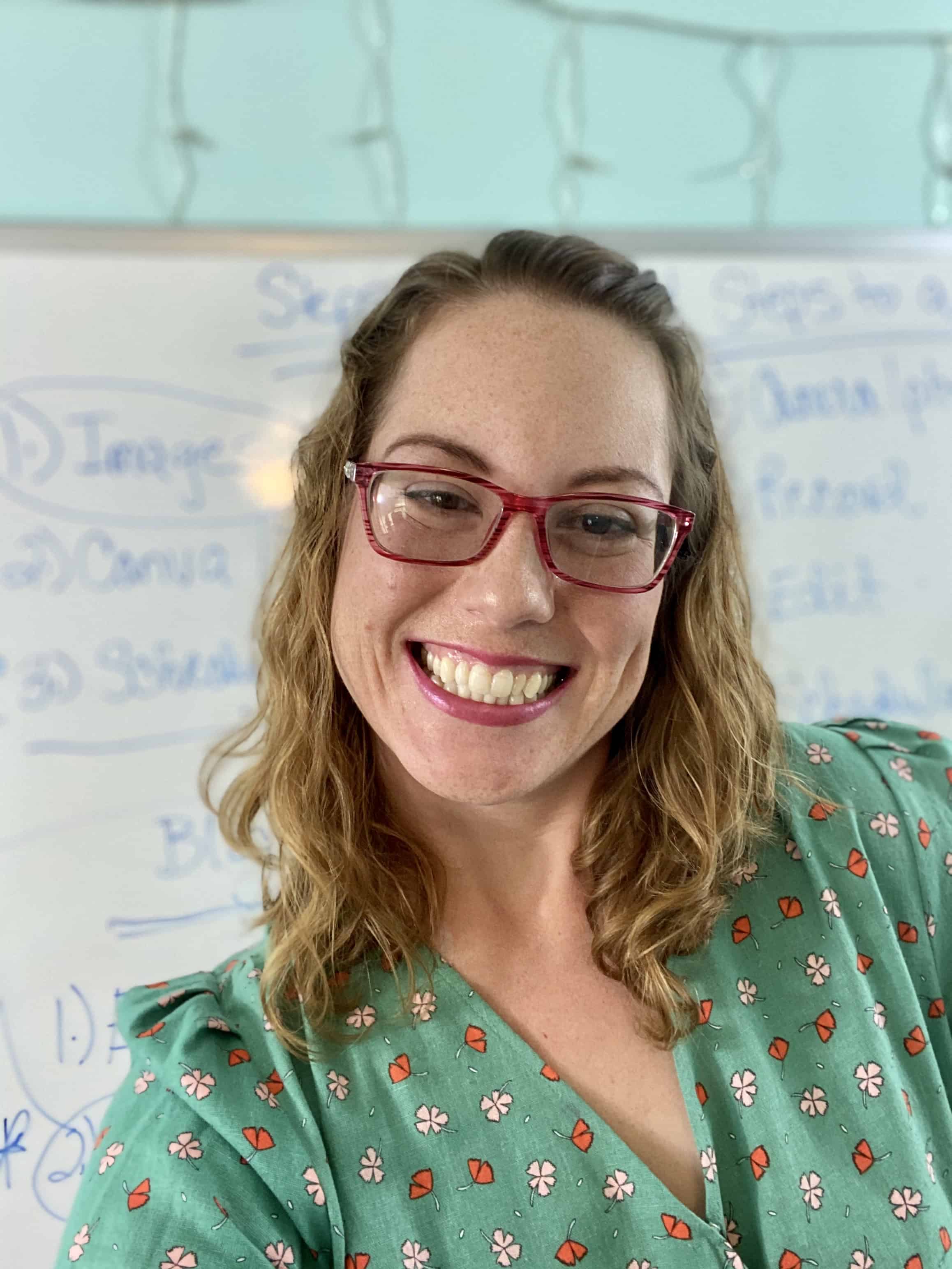 Smiling woman with curly light brown hair, wearing red glasses and a green floral top, with a whiteboard in the background.