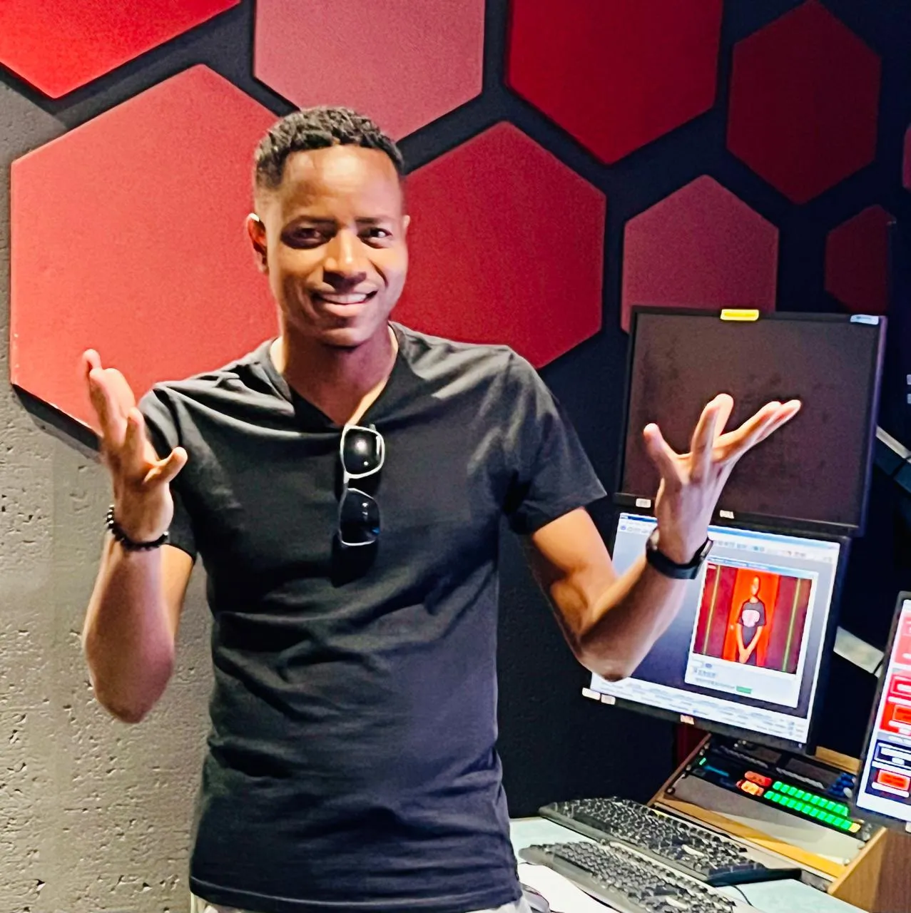 Man smiling with arms raised in a room with red hexagonal wall panels and computer monitors.