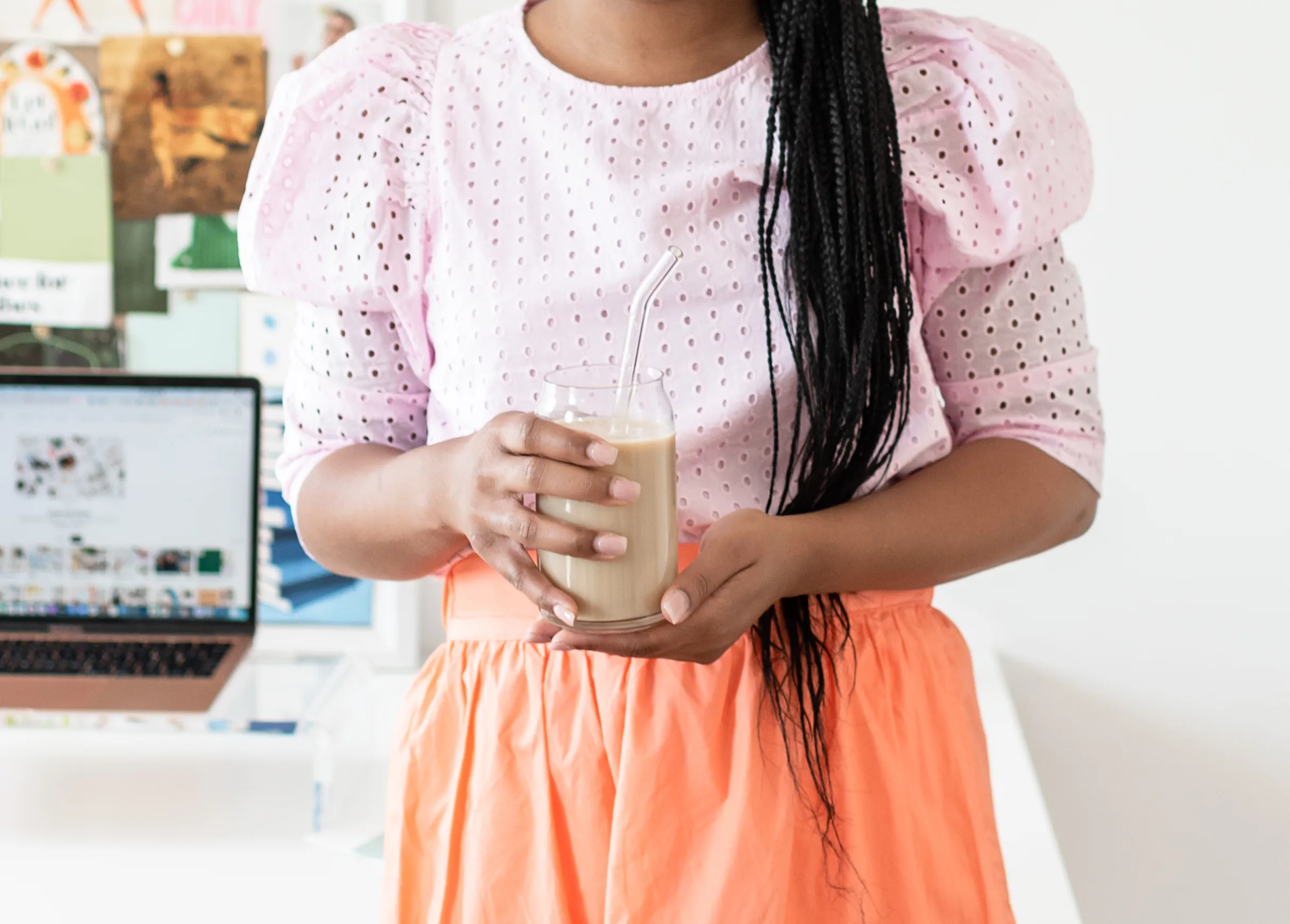 Person in pink eyelet blouse and orange skirt holding a glass of iced coffee with a straw in a bright workspace.