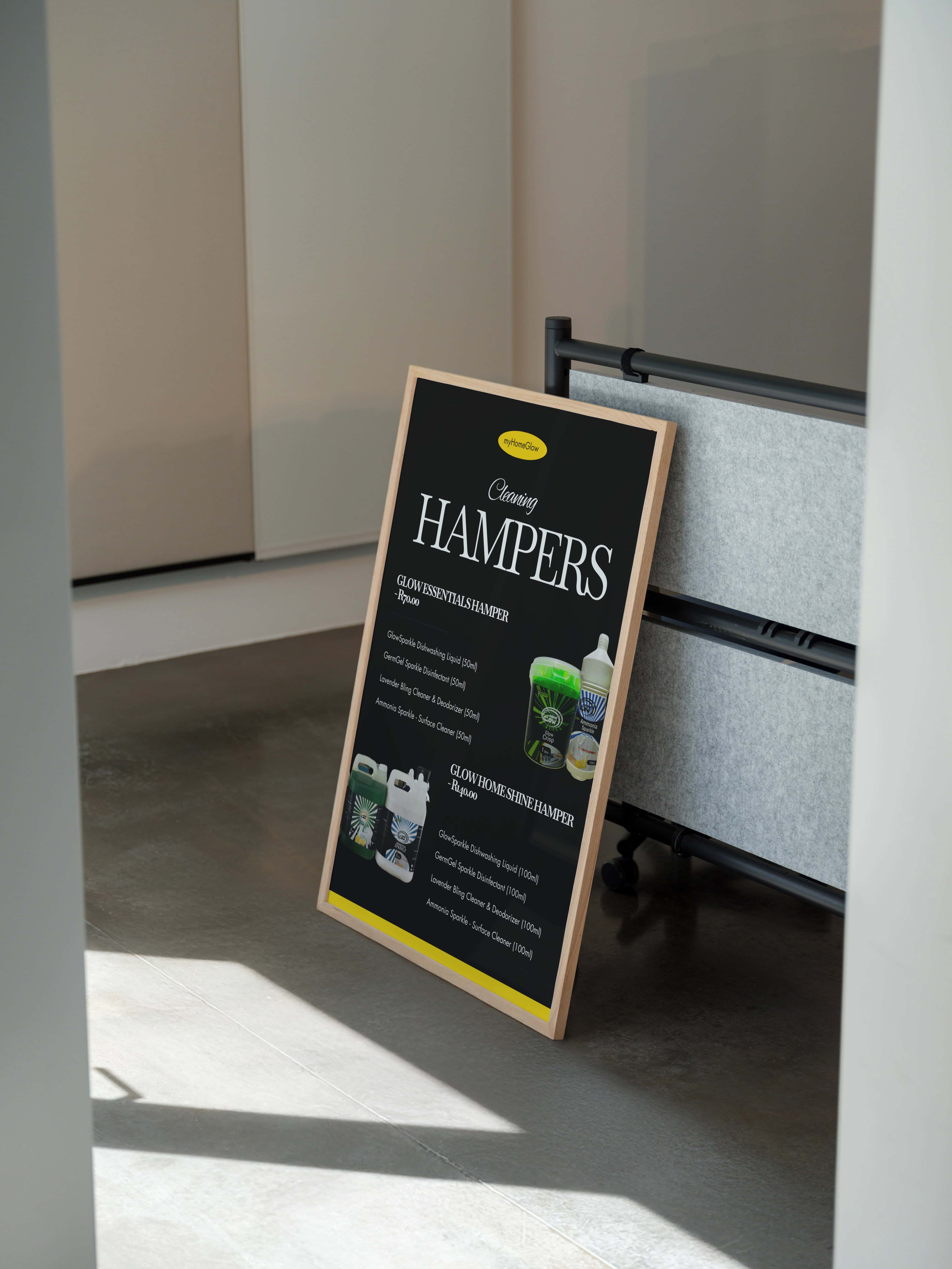 Framed blackboard sign leaning against a gray partition, displaying a menu of cleaning hamper products with images of cleaning bottles and containers.