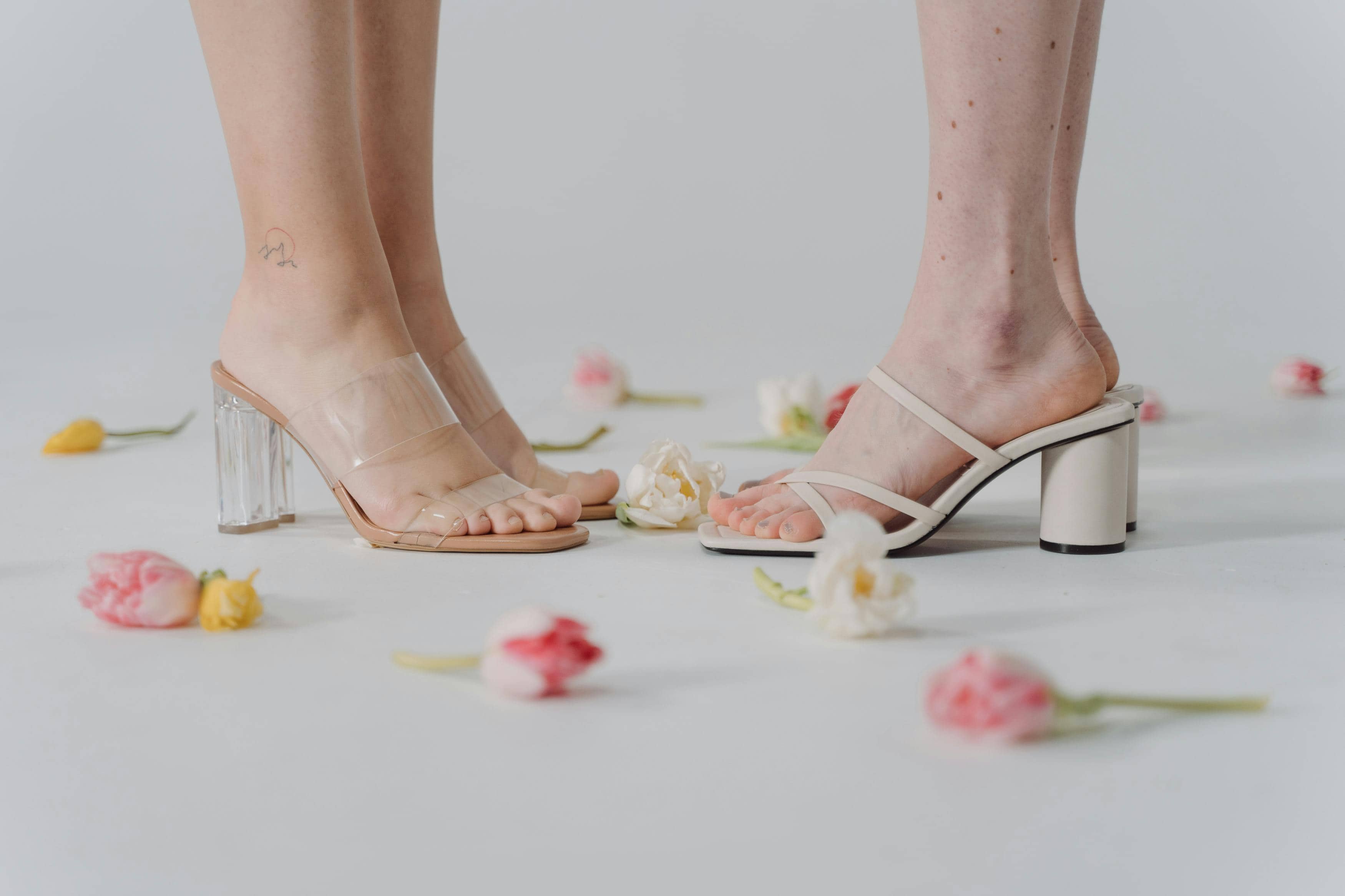 Two women wearing beige heeled sandals standing on a white floor scattered with pink, yellow, and white flowers.