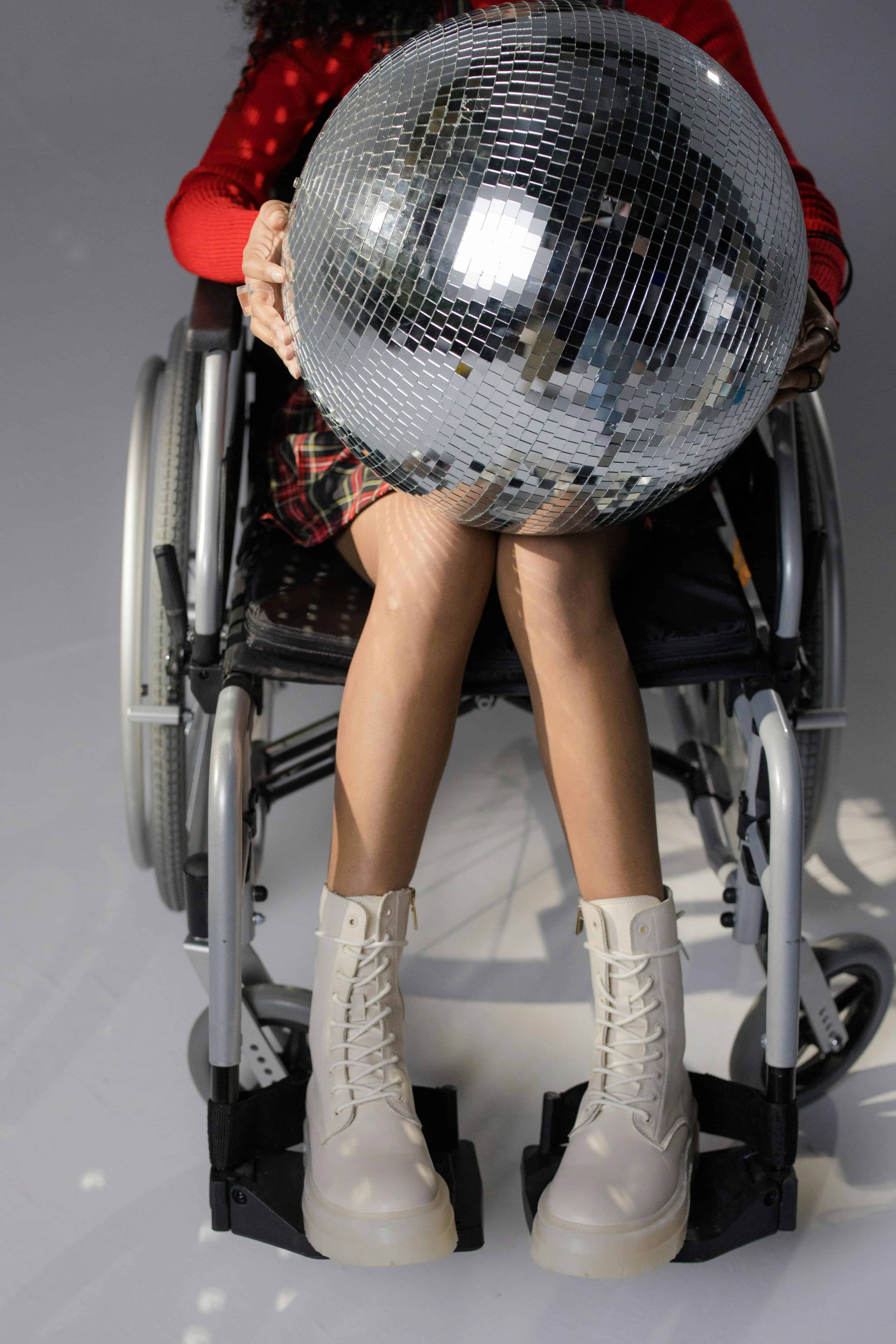 Person in a wheelchair holding a large disco ball, wearing white lace-up boots and a red sweater.