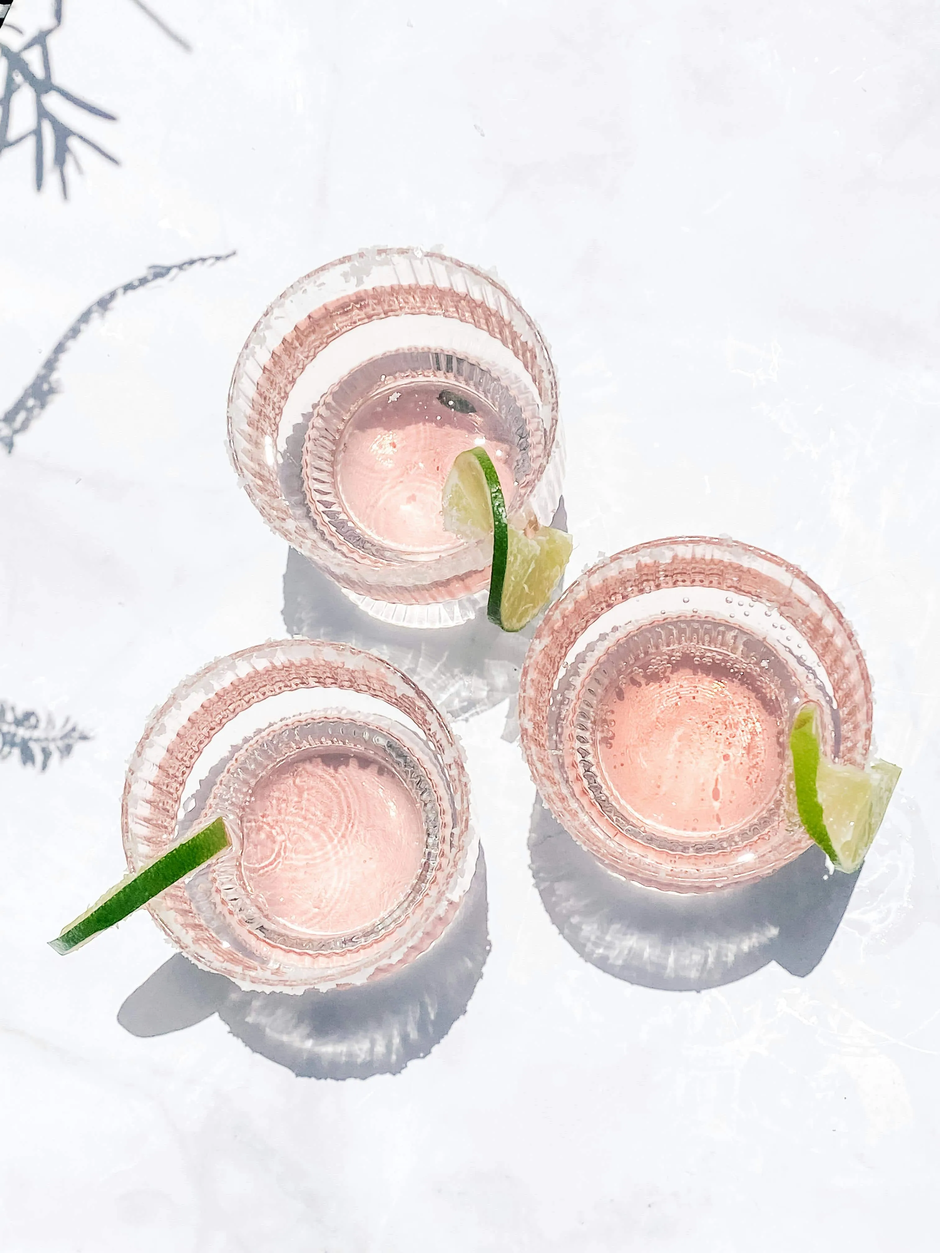 Top view of three pink cocktails in clear glasses, each garnished with a lime wedge on a white surface.
