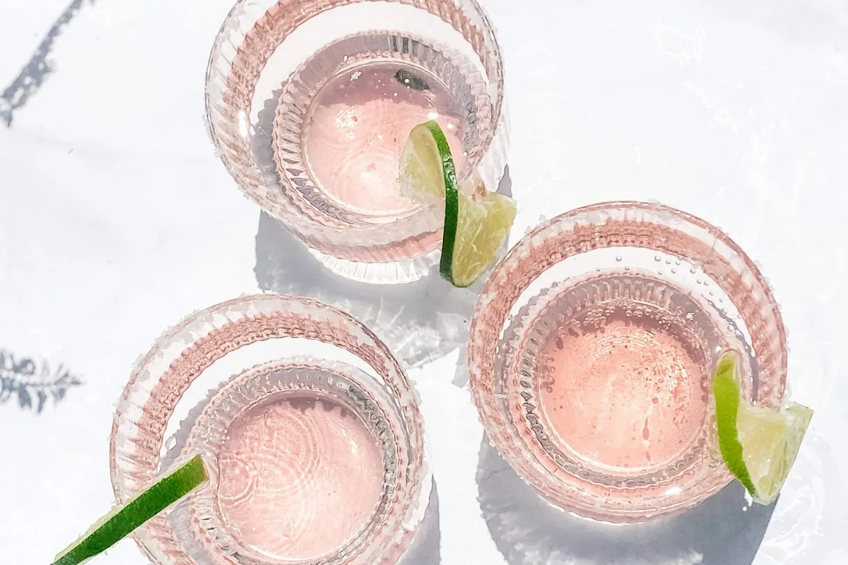 Three glasses with pink sparkling drinks, each garnished with a lime wedge, on a white surface.