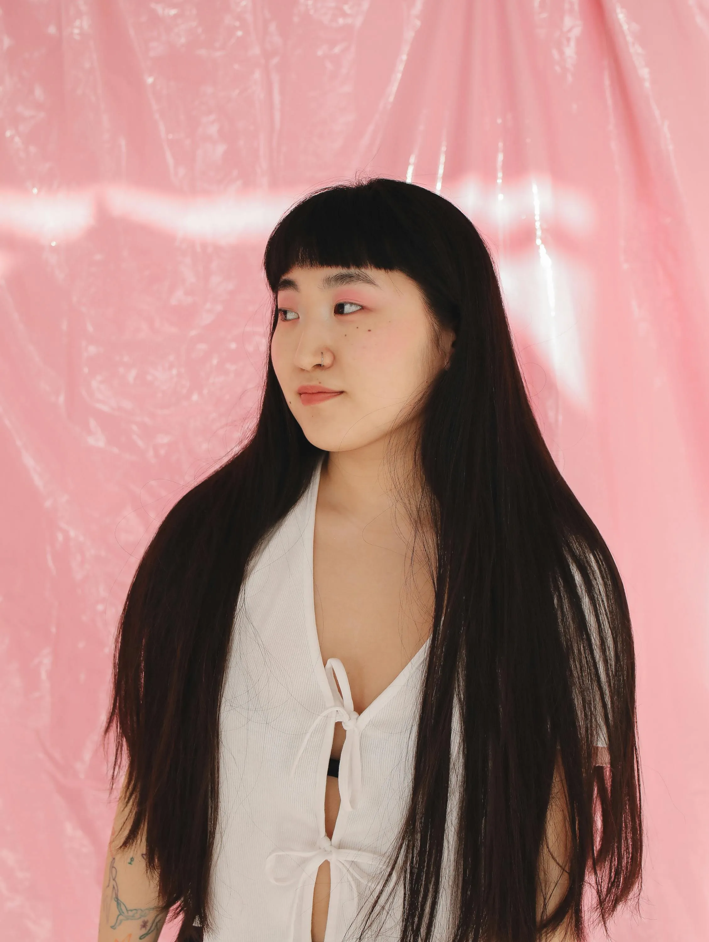 Young woman with long black hair and nose ring wearing a white top, standing against a pink textured background.