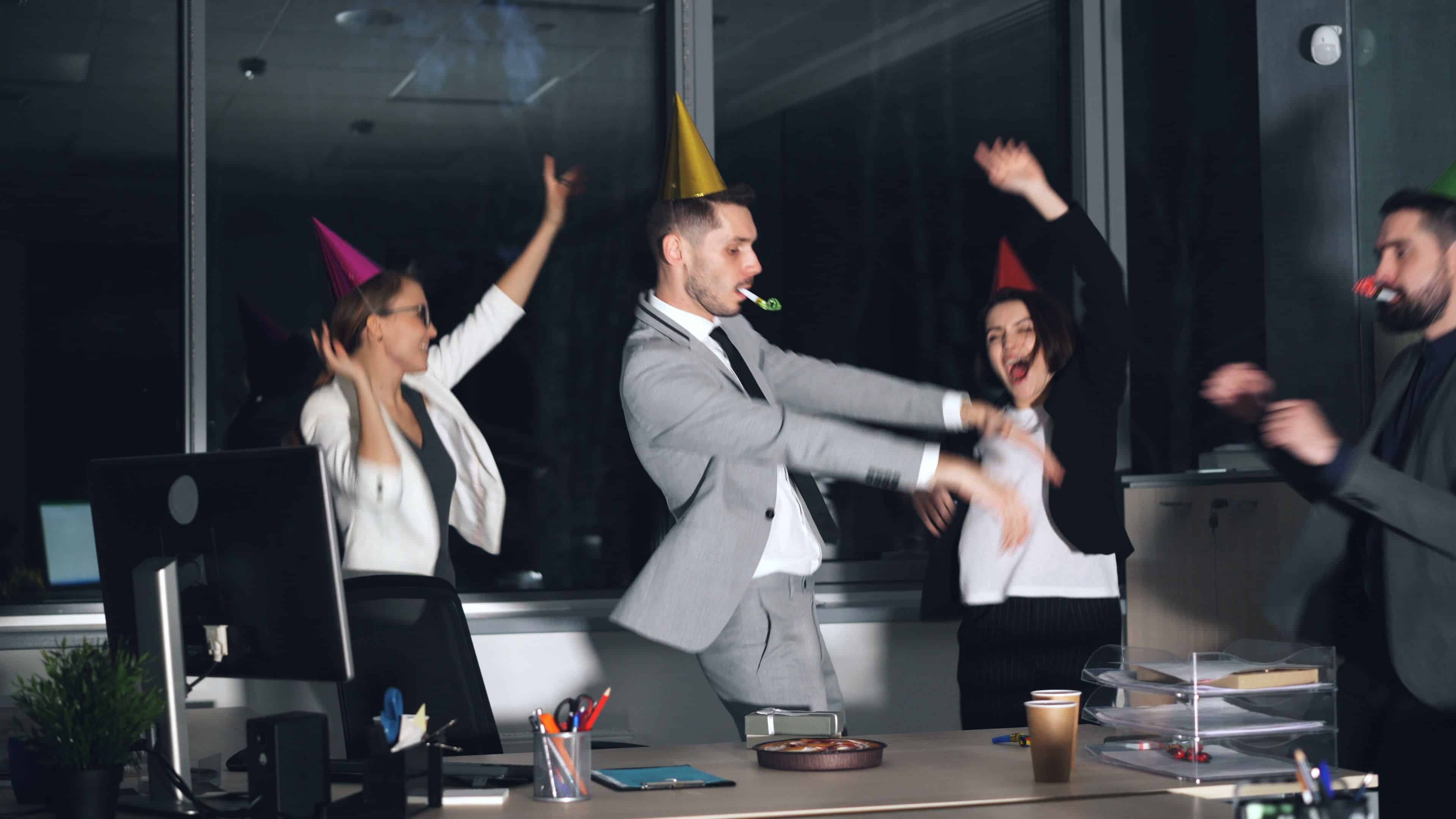 Four coworkers wearing party hats celebrating and dancing in an office after hours.