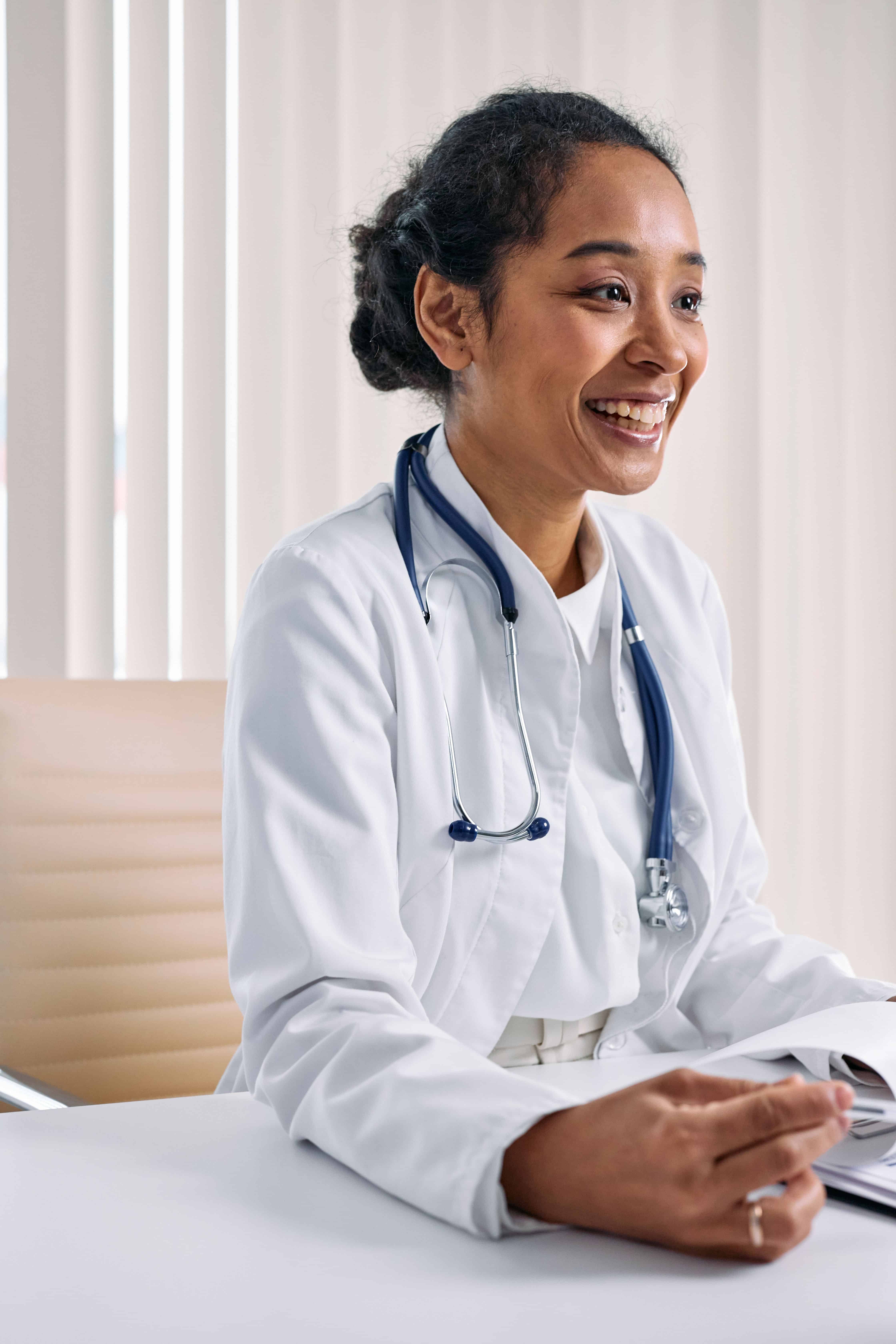 Smiling female doctor in white coat with a stethoscope around her neck, sitting at a desk.