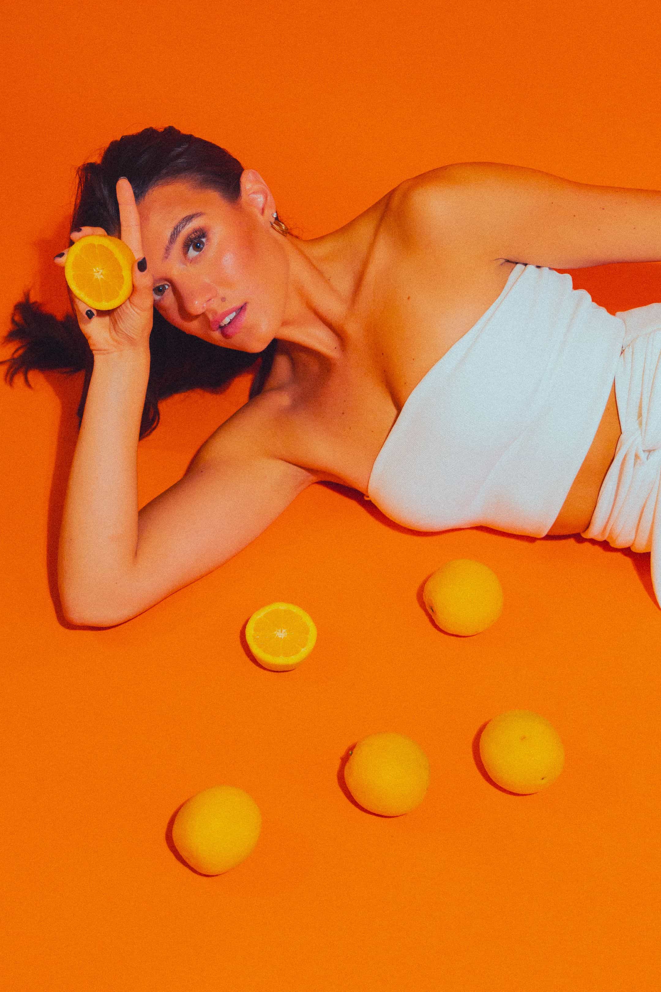 A woman with dark hair lies on an orange background wearing a white tube top, holding half an orange near her face with five whole and one half orange scattered around her.