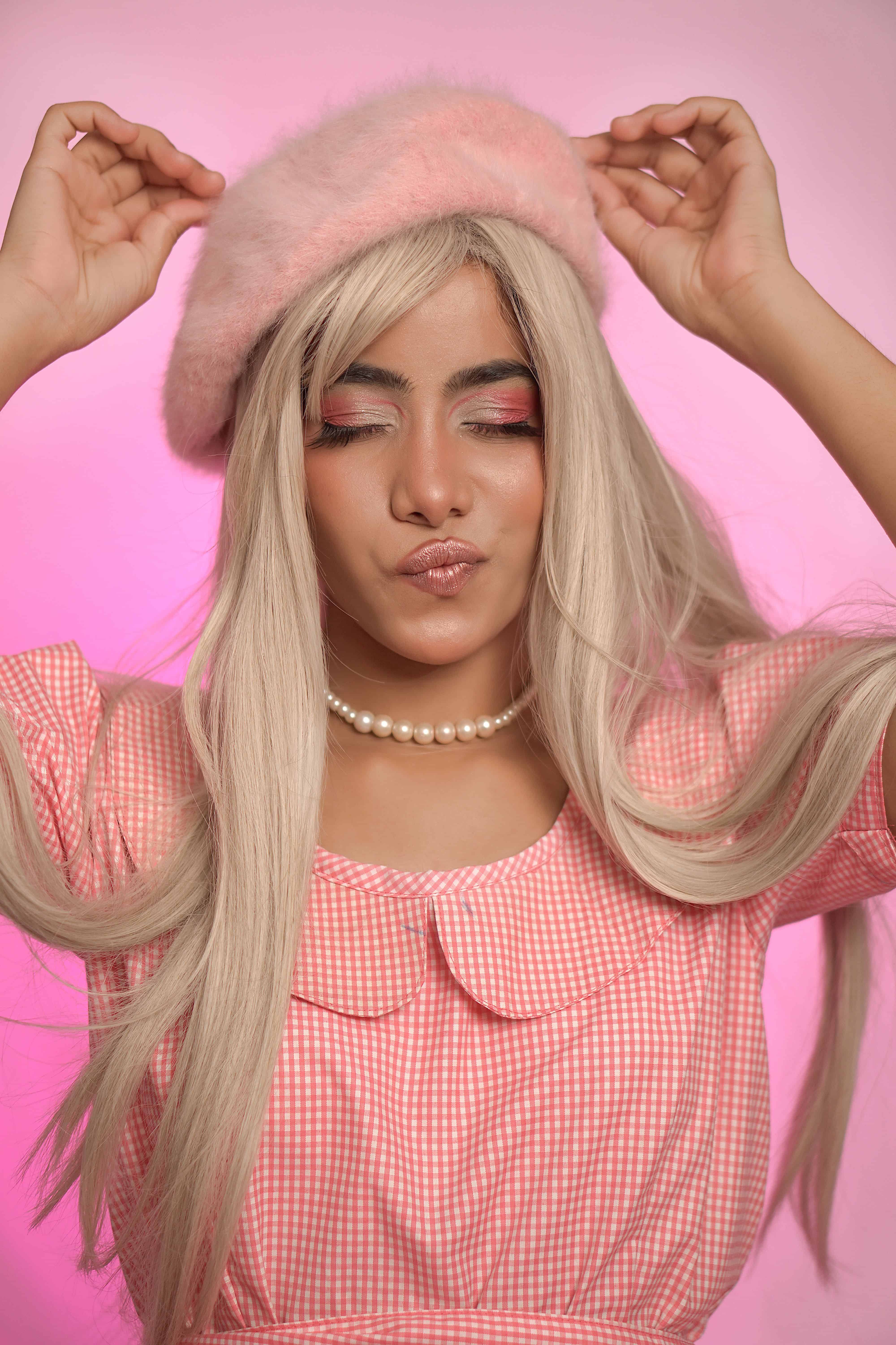 Woman with long blonde hair wearing a pink fuzzy beret, pearl necklace, and checkered pink dress.