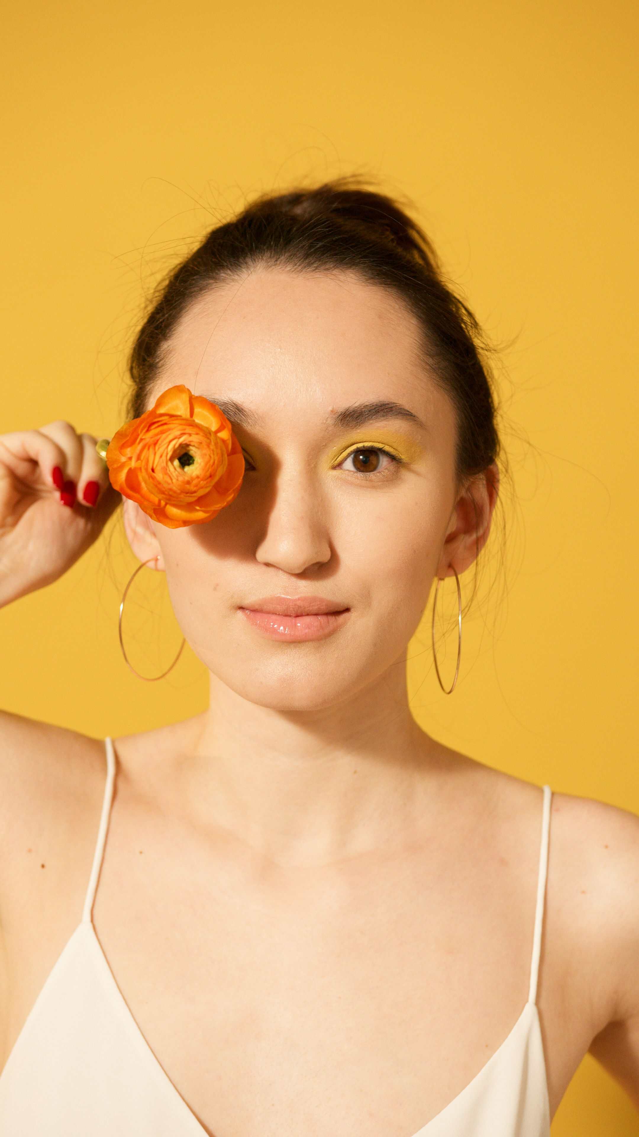 Woman with large hoop earrings holding an orange flower over one eye against a yellow background.