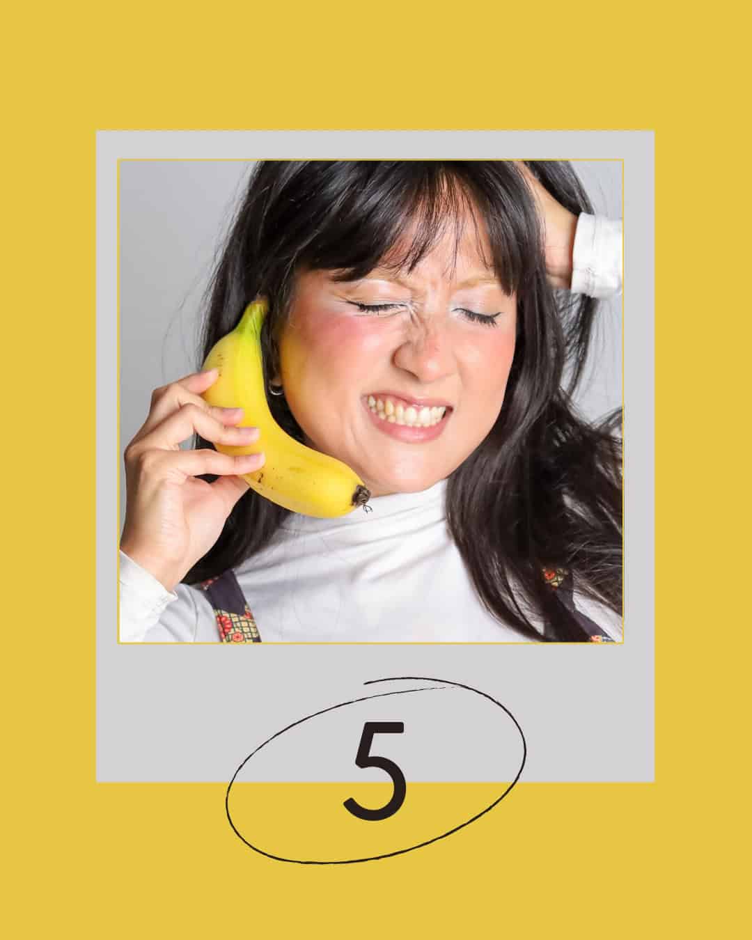 Woman with dark hair holds a banana like a phone to her ear, eyes closed and grimacing.