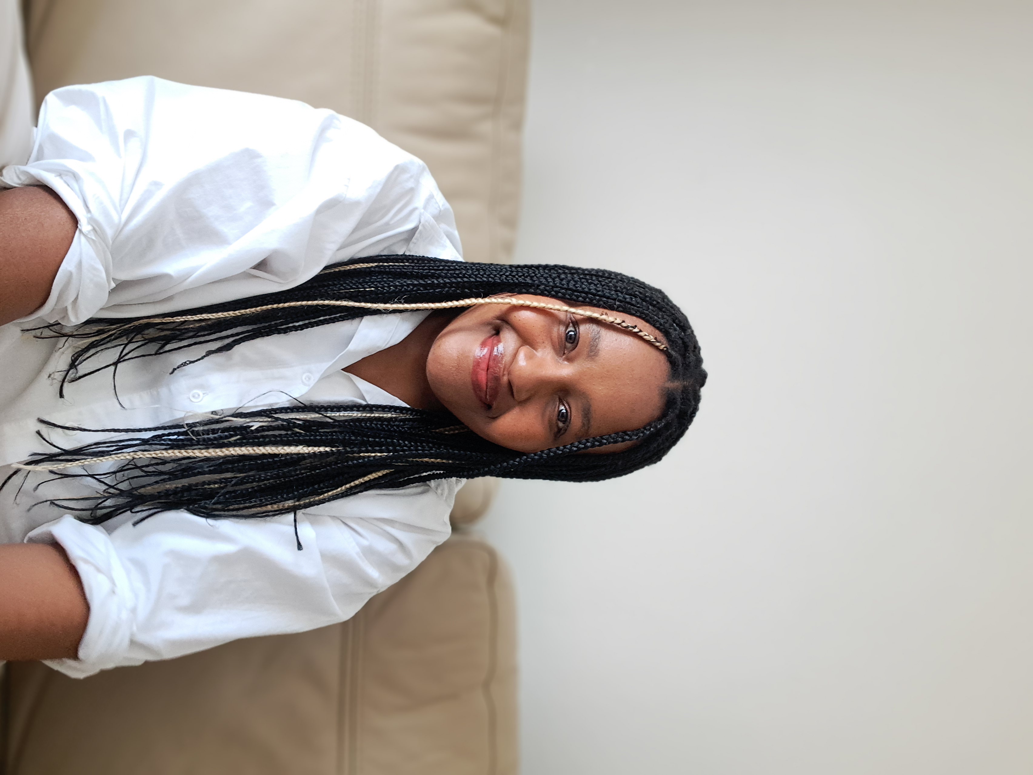 Smiling woman with black and blonde braids wearing glasses and a white shirt sitting on a beige sofa.