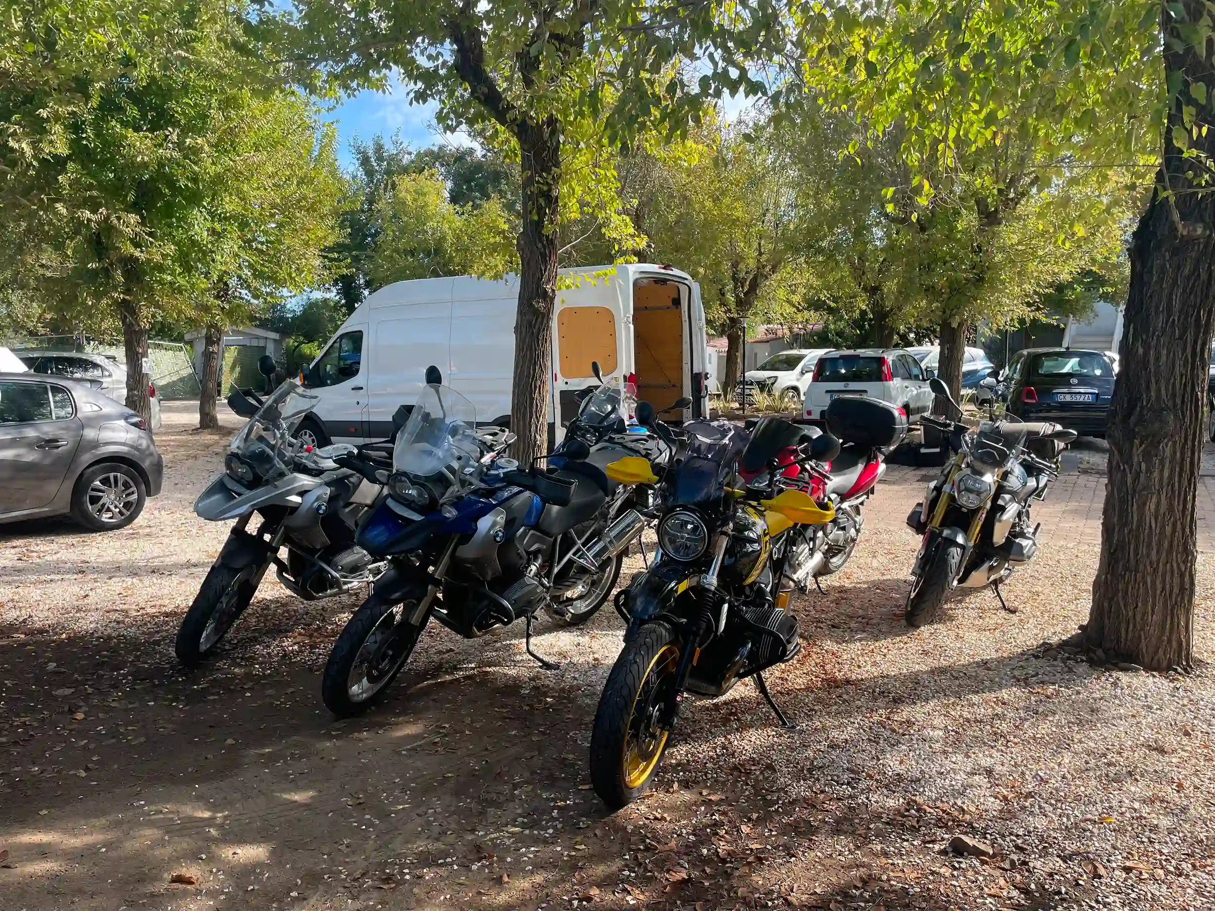 6 Motorräder stehen zwischen den Bäumen auf einem Parkplatz.